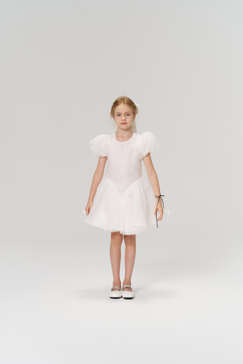 A young girl faces forward in the KIKI and JOJO Ballerina Whisper Dress—a white tulle mini dress with puffed sleeves—paired with white shoes, set against a plain light background.