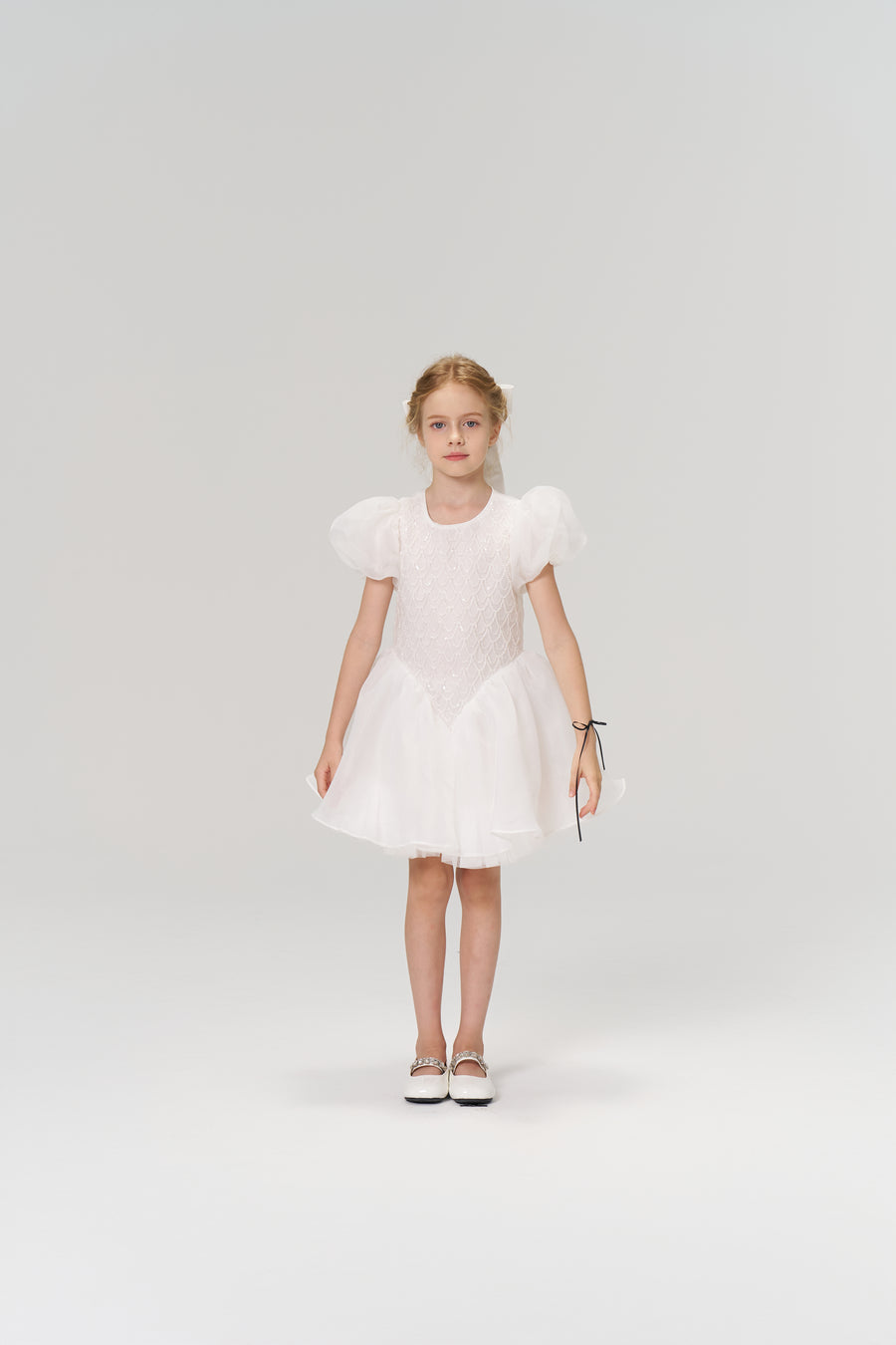 A young girl faces forward in the KIKI and JOJO Ballerina Whisper Dress—a white tulle mini dress with puffed sleeves—paired with white shoes, set against a plain light background.