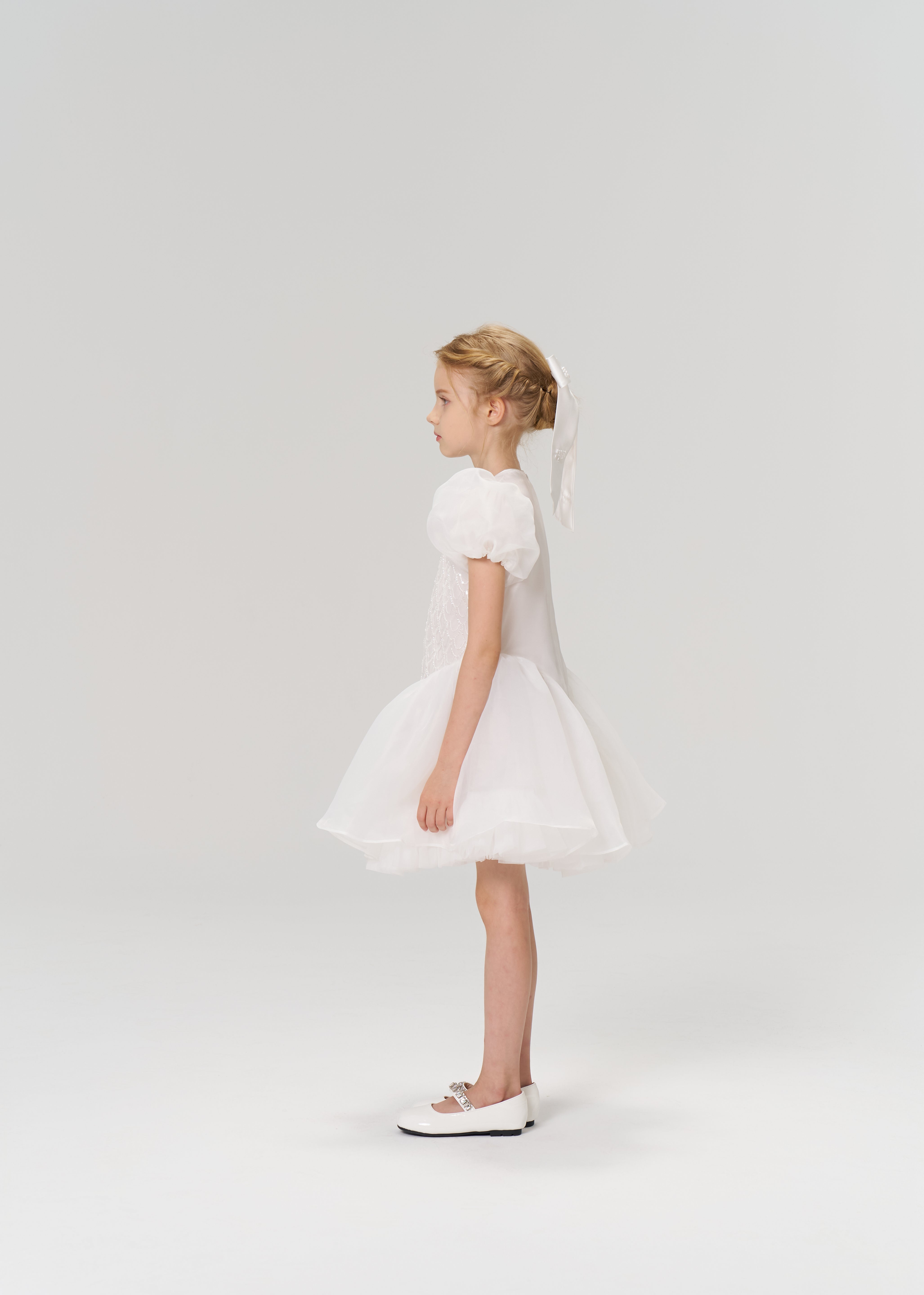 A young girl in profile wears the KIKI and JOJO Ballerina Whisper Dress—a white tulle mini dress with puffed sleeves—paired with white shoes and a ribbon in her braided hair, set against a plain light background.
