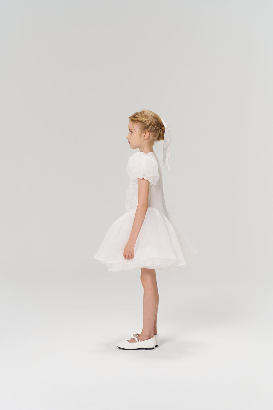 A young girl in profile wears the KIKI and JOJO Ballerina Whisper Dress—a white tulle mini dress with puffed sleeves—paired with white shoes and a ribbon in her braided hair, set against a plain light background.