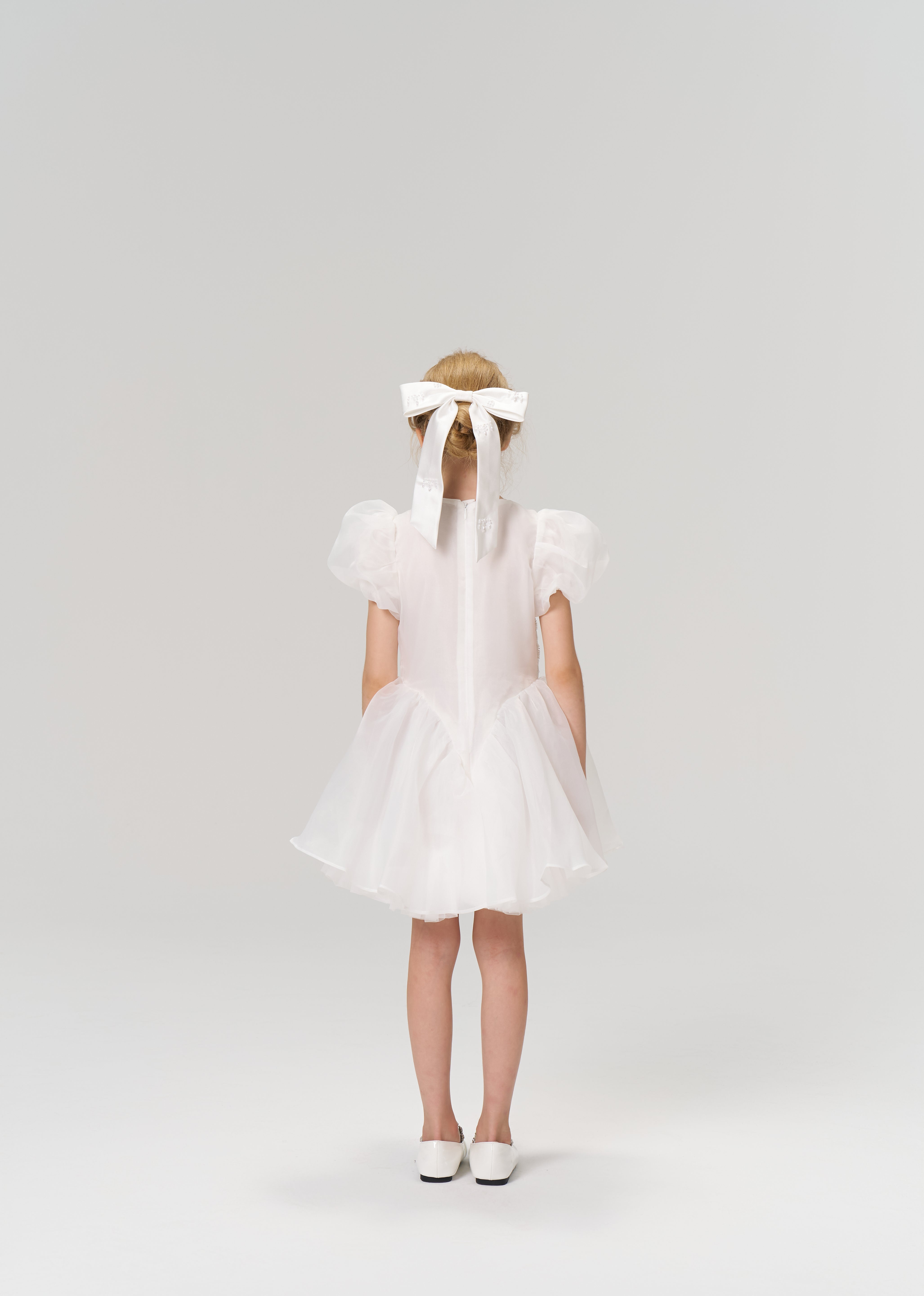A young girl faces away, wearing the KIKI and JOJO Ballerina Whisper Dress—white tulle mini dress with puffed sleeves—paired with white shoes and a large white hair bow, set against a plain background.