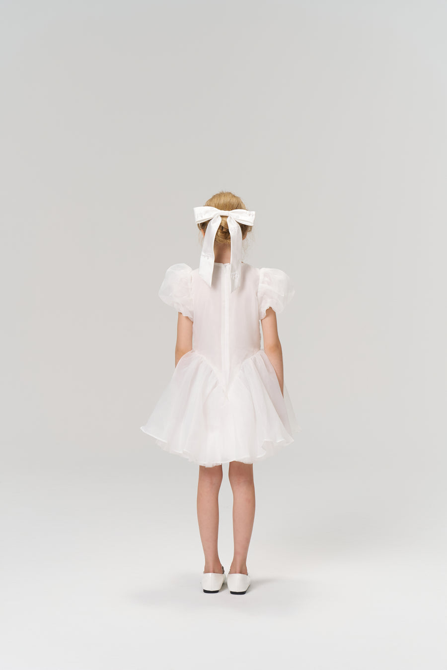 A young girl faces away, wearing the KIKI and JOJO Ballerina Whisper Dress—white tulle mini dress with puffed sleeves—paired with white shoes and a large white hair bow, set against a plain background.