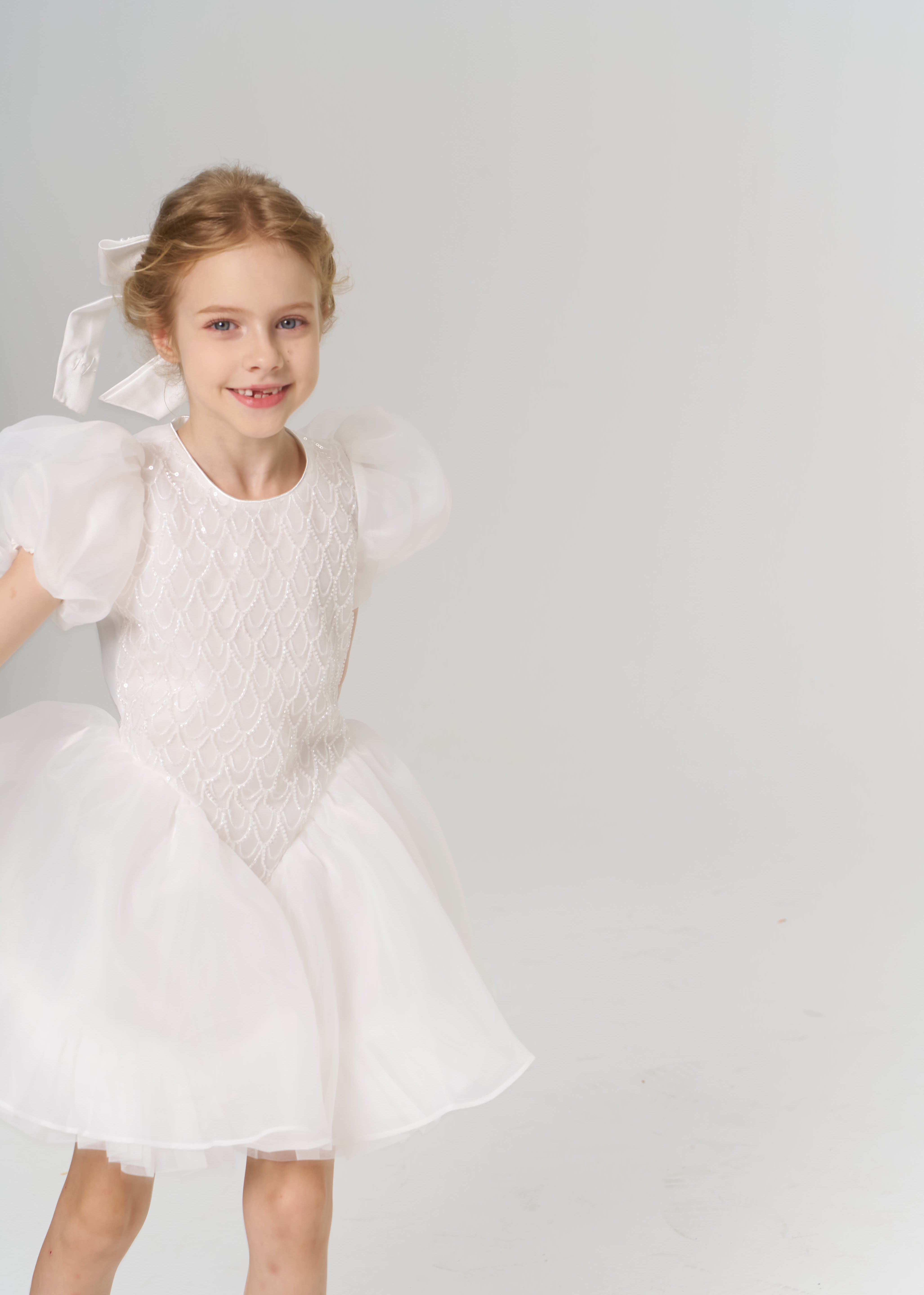 Young girl wearing a white dress with puffed sleeves and a bow in her hair, standing against a plain light background.