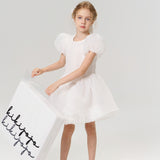 A young girl in a white dress and shoes holds a large KIKI and JOJO Gift Packaging box with elegant black cursive, making it the perfect gift wrap for any special occasion, set against a plain light background.