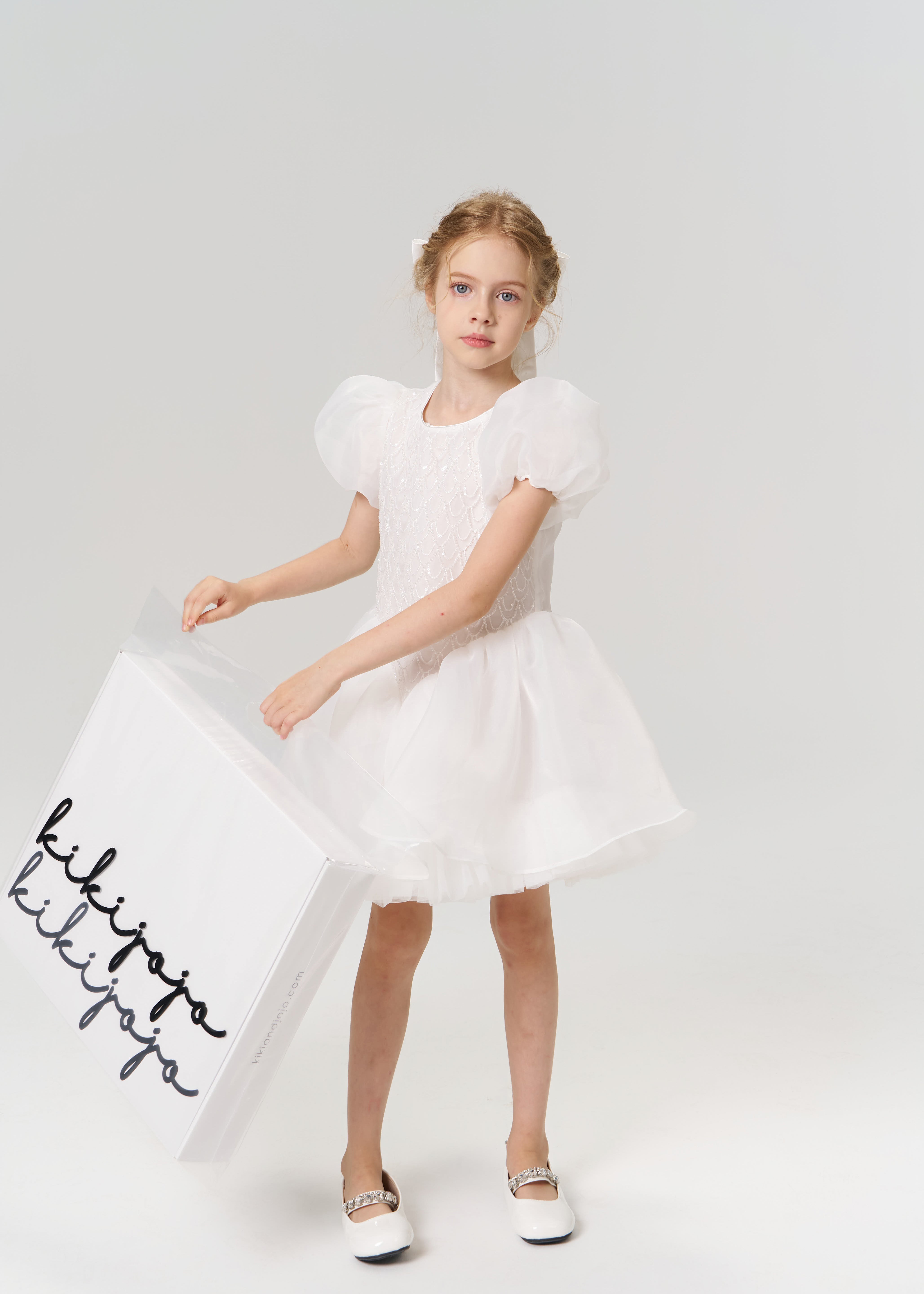 A young girl in a white dress and shoes holds a large KIKI and JOJO Gift Packaging box with elegant black cursive, making it the perfect gift wrap for any special occasion, set against a plain light background.