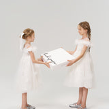 Two young girls in white dresses and silver shoes face each other, holding a large KIKI and JOJO Gift Packaging box—ideal for special moments—against a plain background.