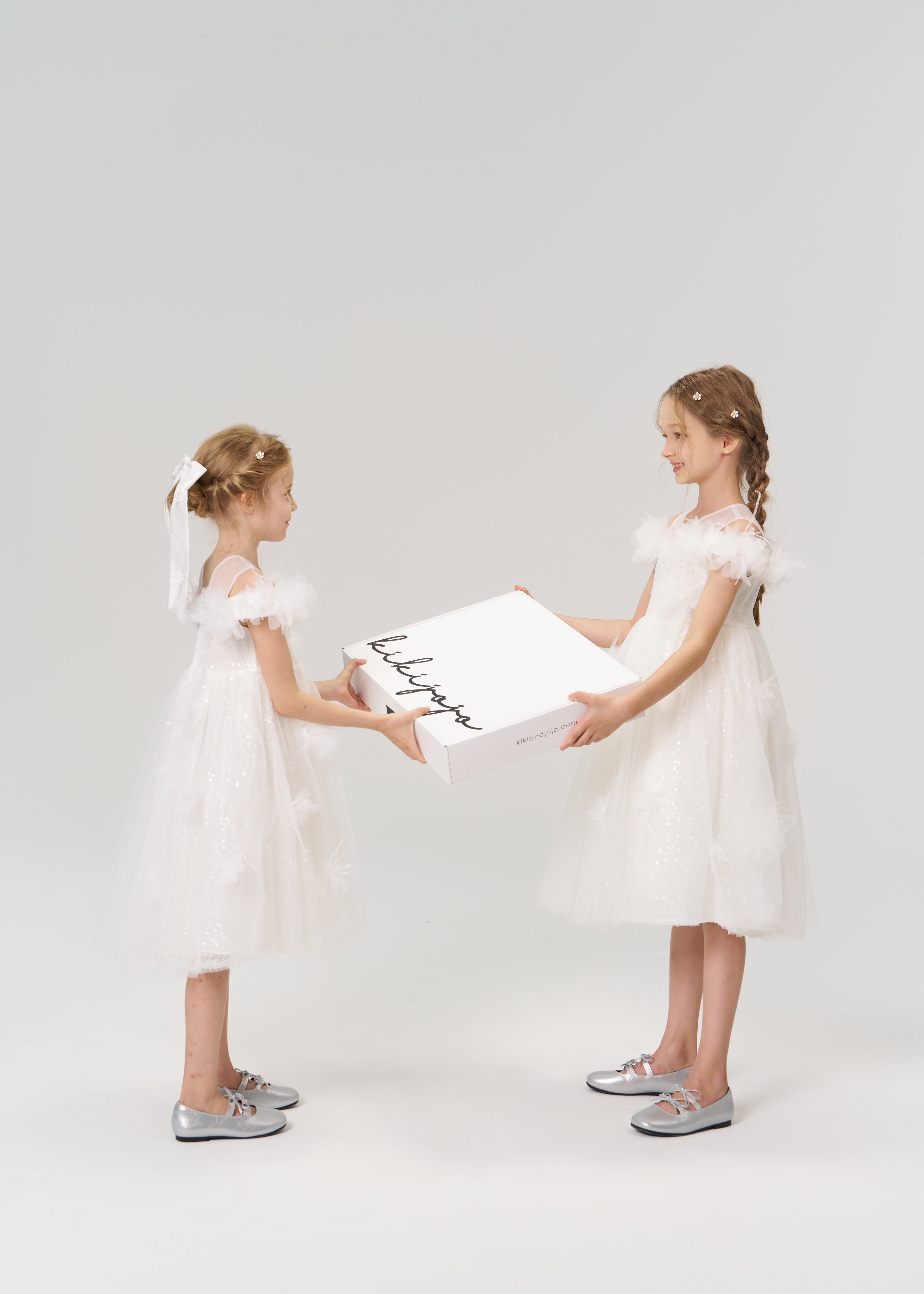 Two young girls in white dresses and silver shoes face each other, holding a large KIKI and JOJO Gift Packaging box—ideal for special moments—against a plain background.