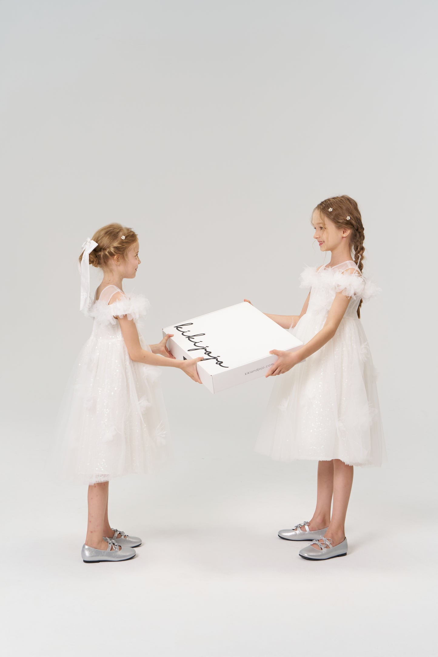 Two young girls in white dresses and silver shoes face each other, holding a large KIKI and JOJO Gift Packaging box—ideal for special moments—against a plain background.