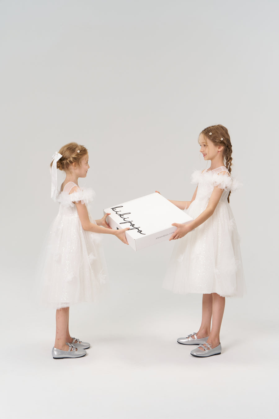 Two young girls in white dresses and silver shoes face each other, holding a large KIKI and JOJO Gift Packaging box—ideal for special moments—against a plain background.