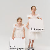 Two young girls in white dresses hold large white boxes labeled "KIKI and JOJO" Gift Packaging, ideal for presenting gifts, against a plain light background.