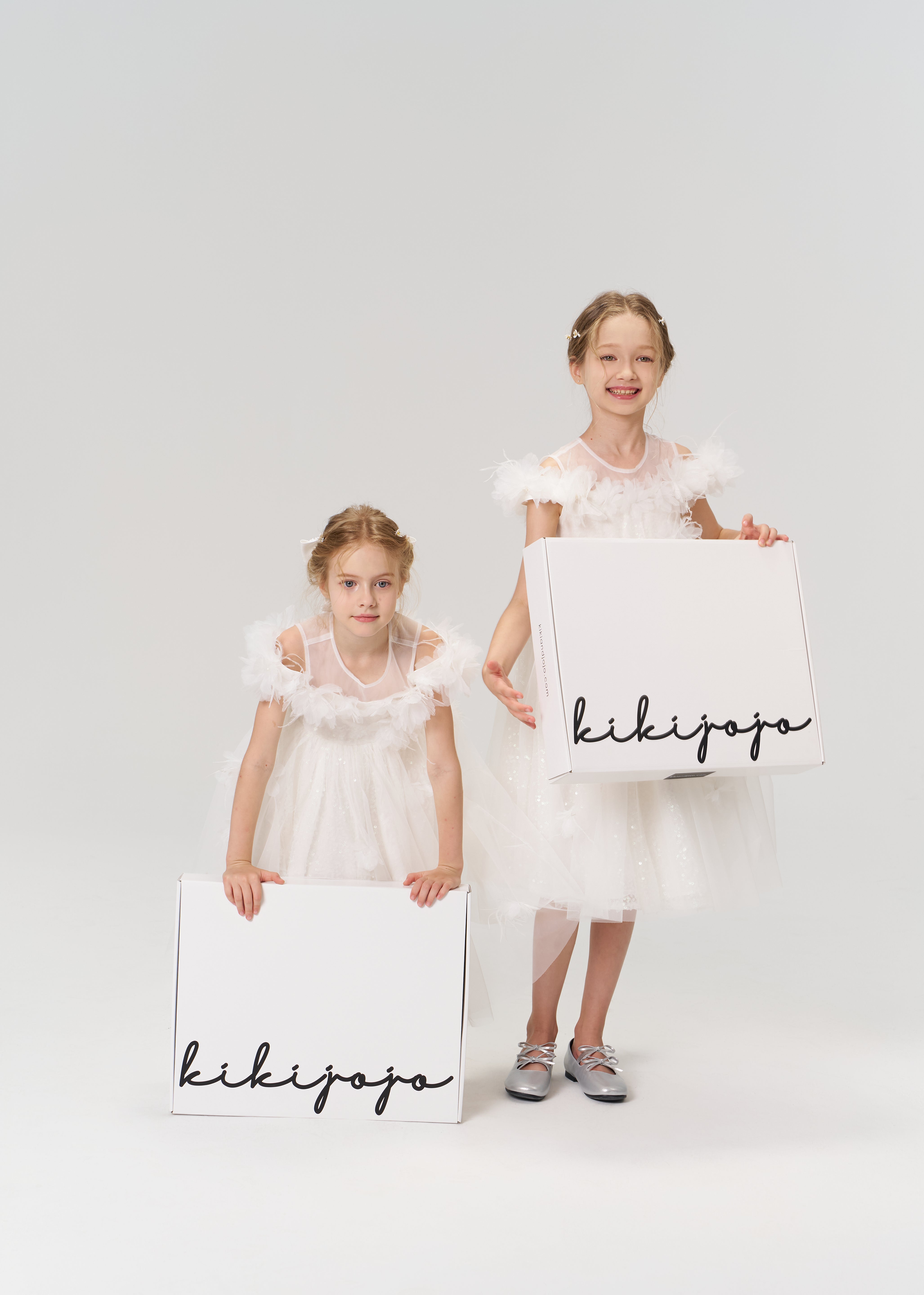 Two young girls in white dresses hold large white boxes labeled "KIKI and JOJO" Gift Packaging, ideal for presenting gifts, against a plain light background.
