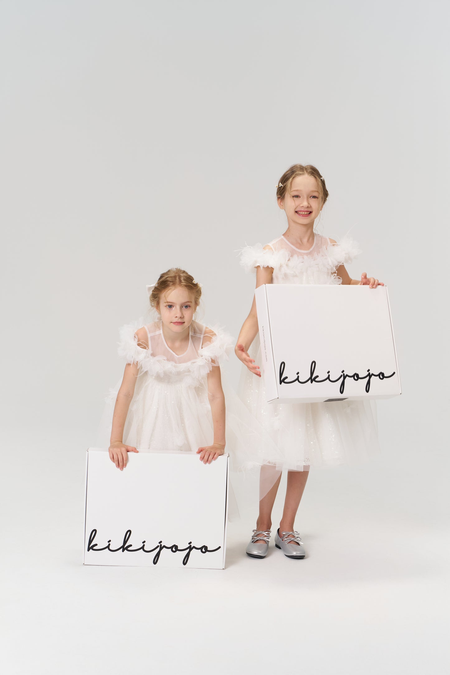 Two young girls in white dresses hold large white boxes labeled "KIKI and JOJO" Gift Packaging, ideal for presenting gifts, against a plain light background.