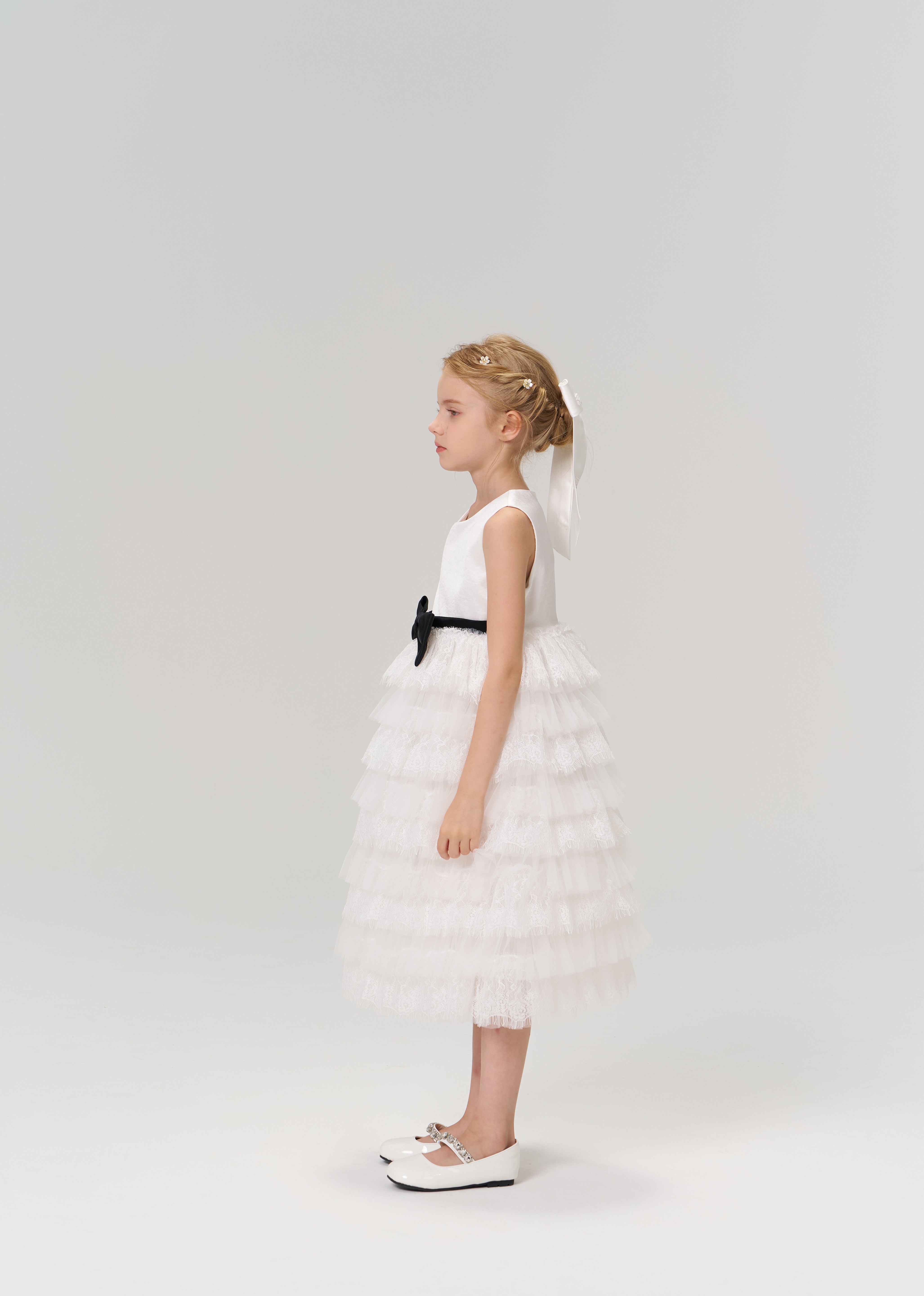 A young girl stands in profile against a plain light background, wearing the KIKI and JOJO Monochrome Whisper Dress with ruffled layers, a black belt, and white shoes.