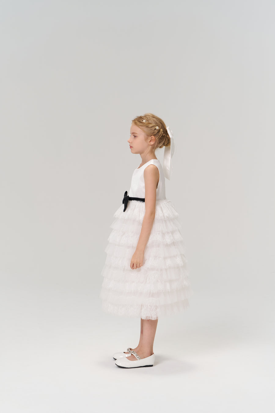 A young girl stands in profile against a plain light background, wearing the KIKI and JOJO Monochrome Whisper Dress with ruffled layers, a black belt, and white shoes.