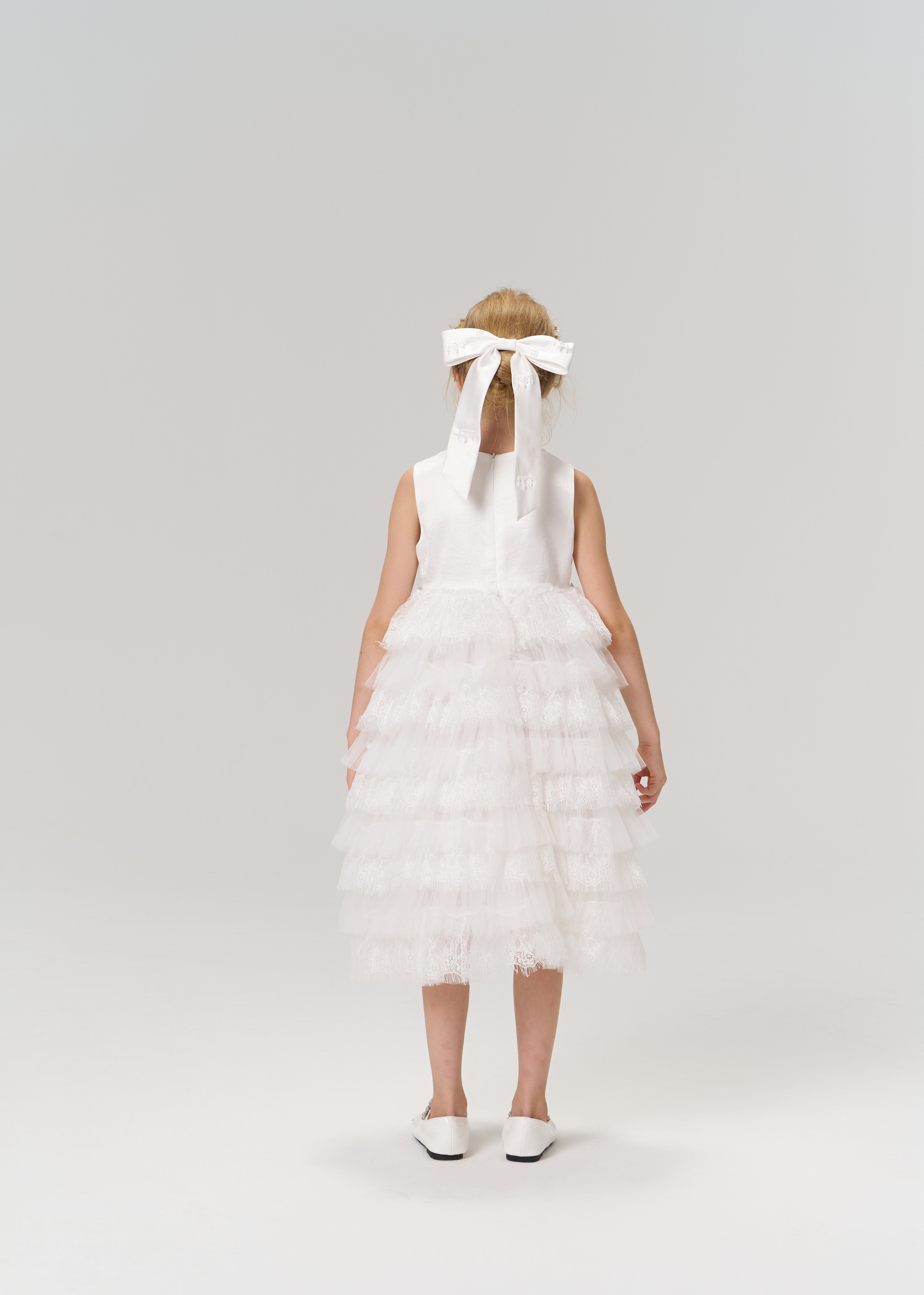 A young girl faces away, wearing the KIKI and JOJO Monochrome Whisper Dress—a white, tiered tulle cake skirt—paired with white shoes and a large white bow in her hair against a plain background.