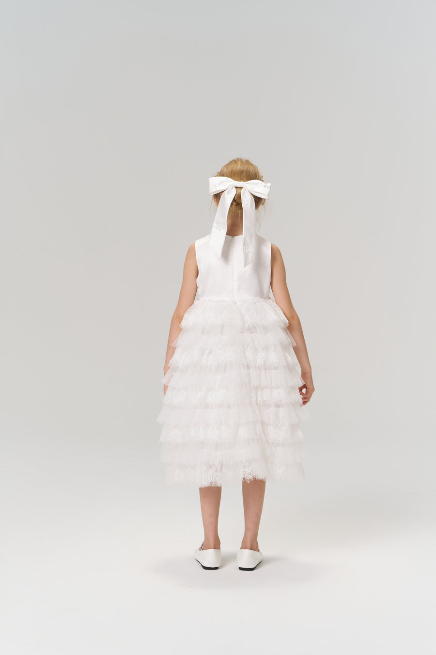 A young girl faces away, wearing the KIKI and JOJO Monochrome Whisper Dress—a white, tiered tulle cake skirt—paired with white shoes and a large white bow in her hair against a plain background.