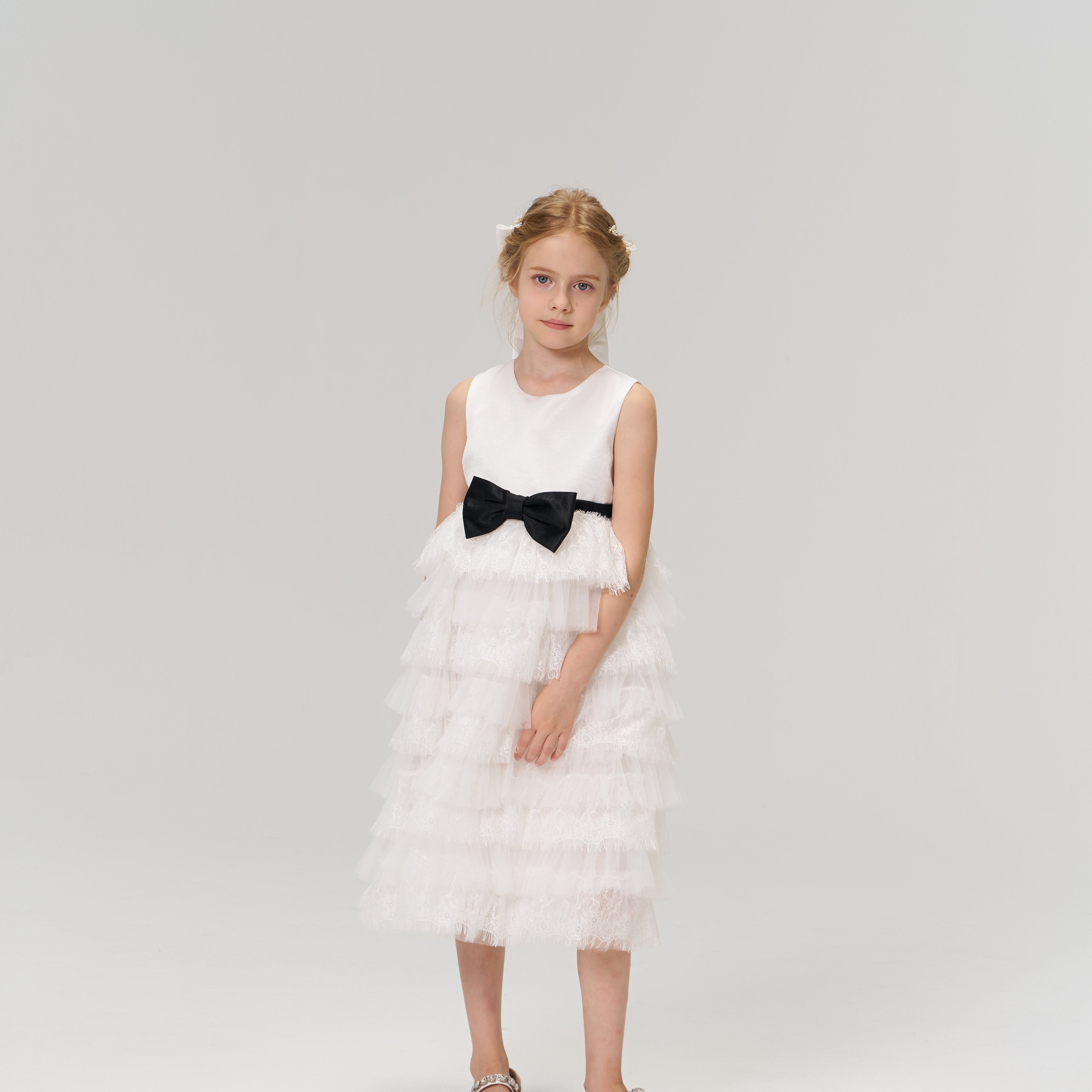 A young girl poses against a plain background in the KIKI and JOJO Monochrome Whisper Dress—a sleeveless, black and white layered dress with ruffles and a black bow—paired with white shoes.