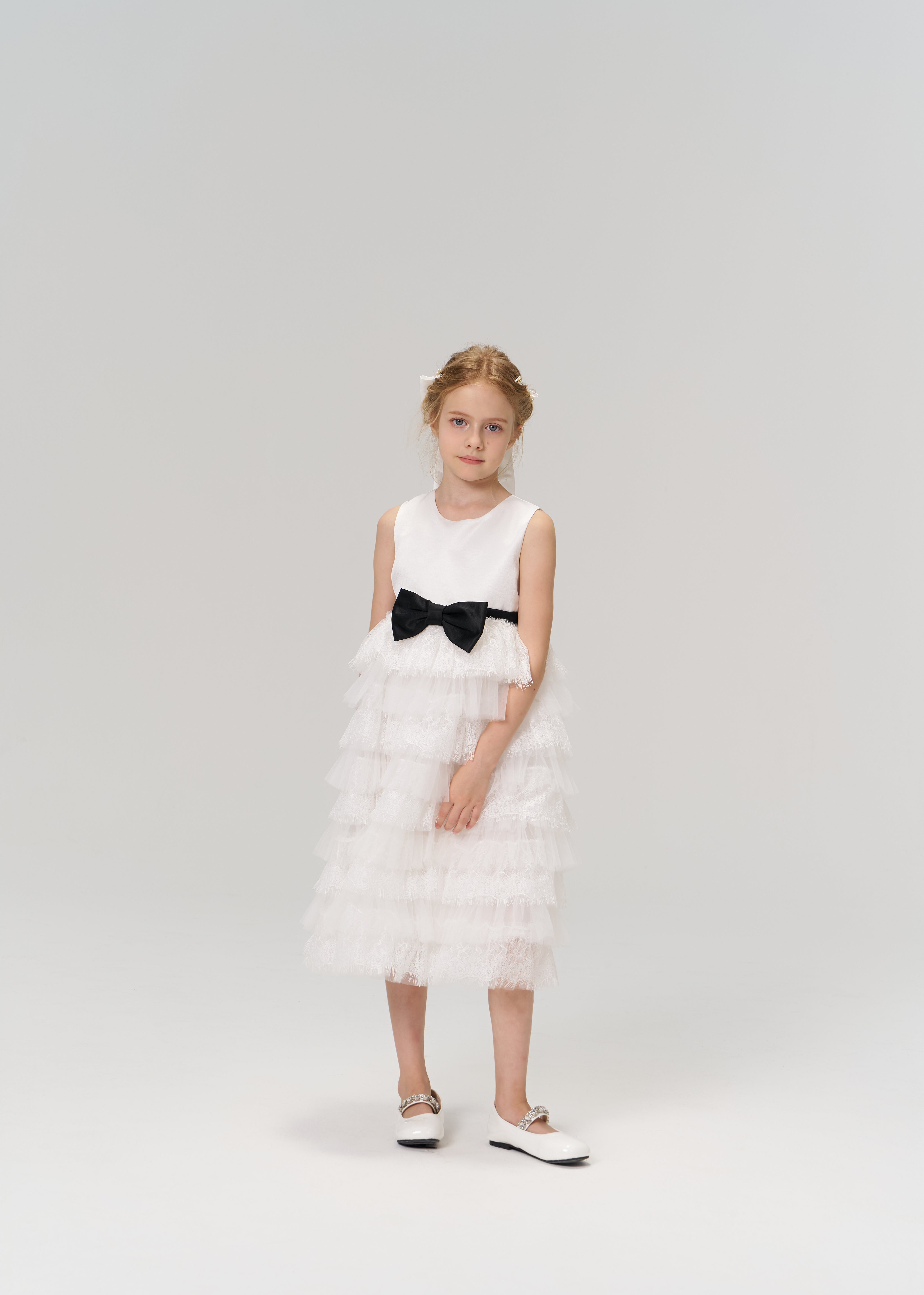 A young girl poses against a plain background in the KIKI and JOJO Monochrome Whisper Dress—a sleeveless, black and white layered dress with ruffles and a black bow—paired with white shoes.