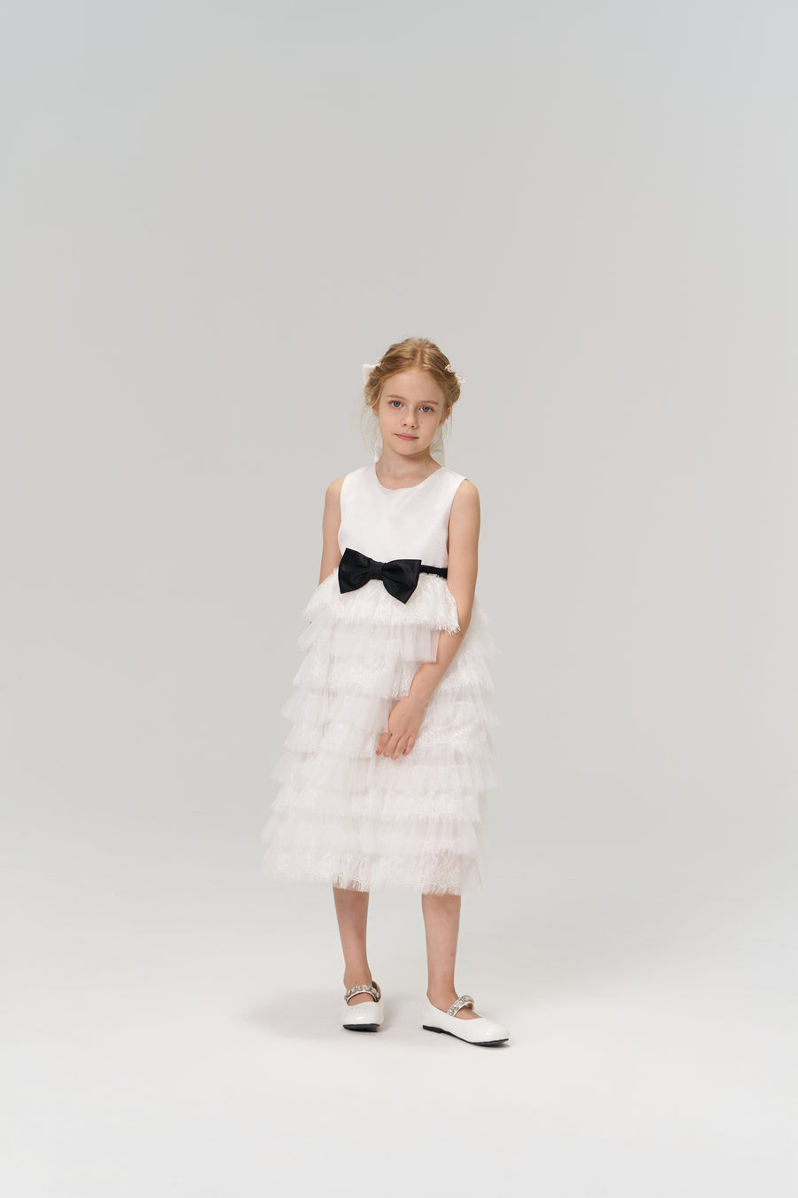 A young girl poses against a plain background in the KIKI and JOJO Monochrome Whisper Dress—a sleeveless, black and white layered dress with ruffles and a black bow—paired with white shoes.