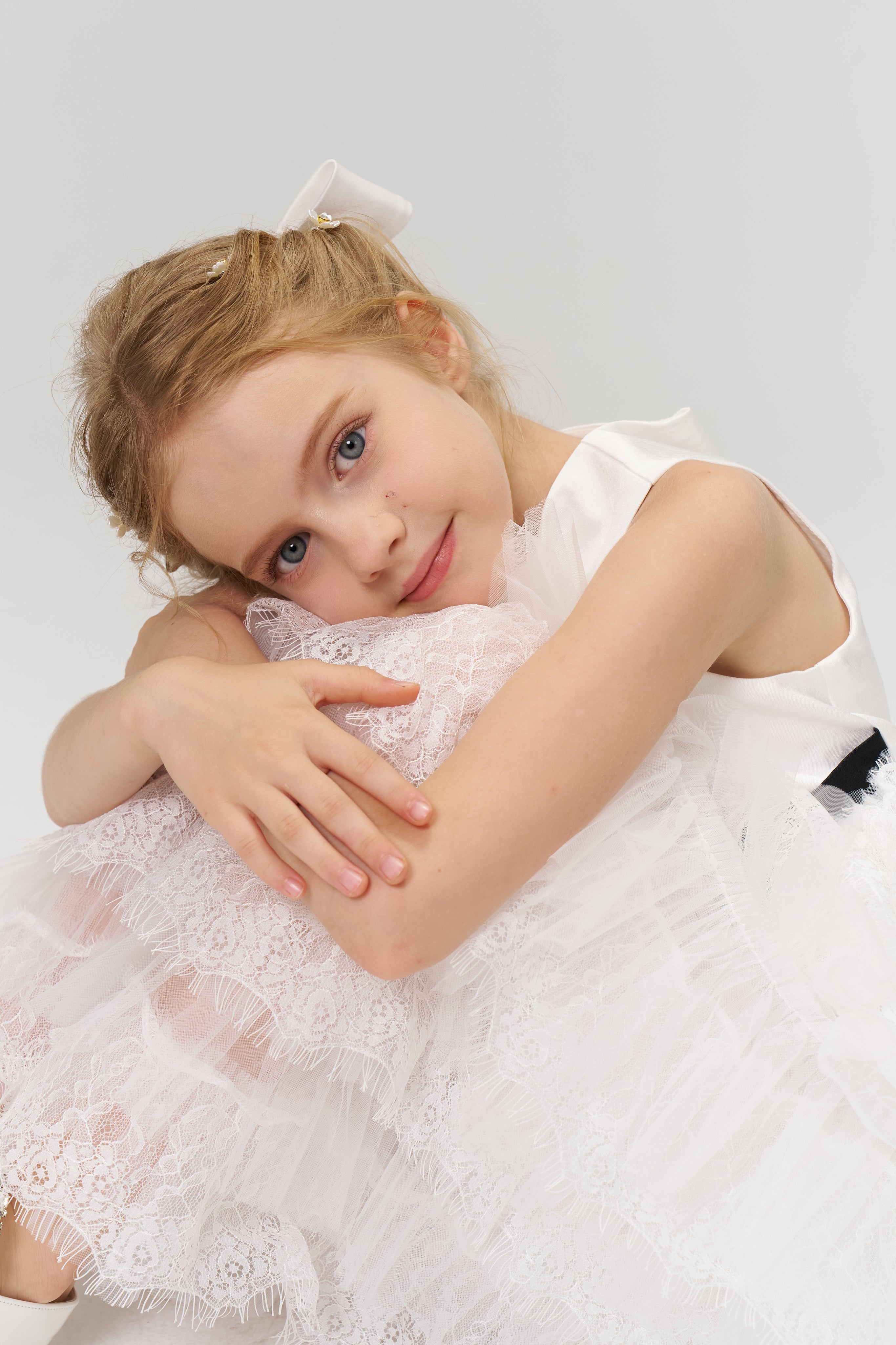 Young girl in a white lace dress sits with her arms wrapped around her knees, looking at the camera with a gentle expression against a plain light background.