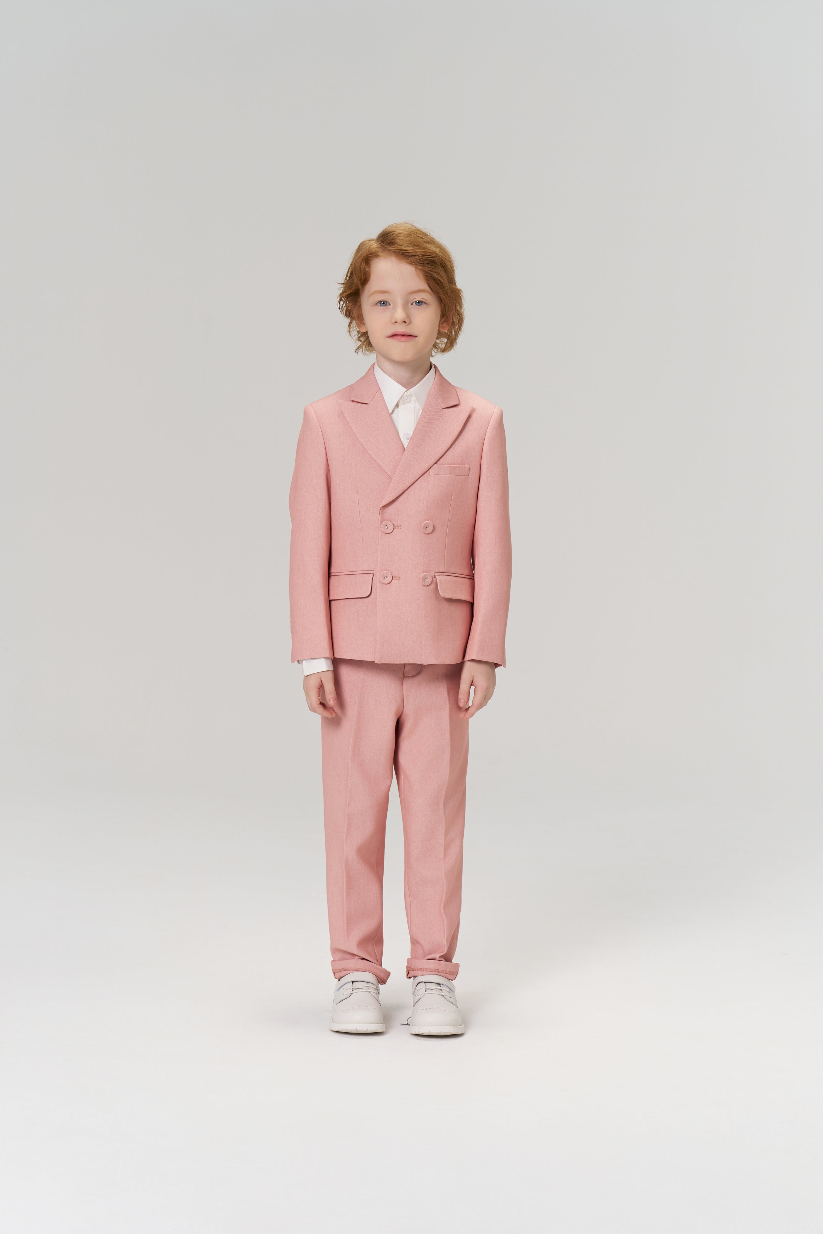 A young child with light brown hair faces forward, wearing KIKI and JOJO’s Blush Reverie Suit Pants with an adjustable waistband, a white shirt, and white sneakers against a plain light background.