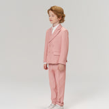 A young boy with light brown hair stands in a studio against a plain light background, wearing KIKI and JOJO’s Blush Reverie Suit Pants with an adjustable waistband, paired with a white shirt and white sneakers.