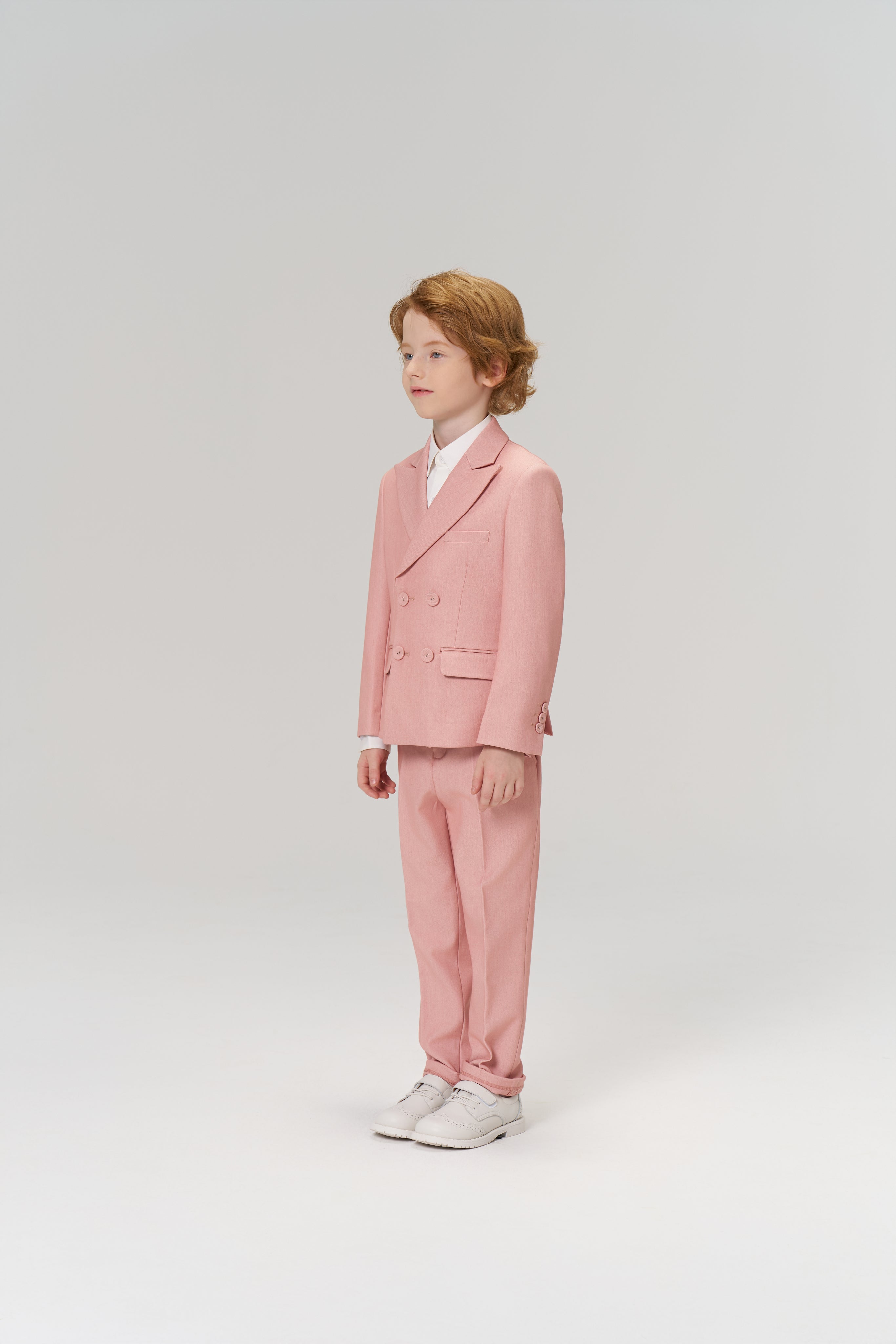 A young boy with light brown hair stands in a studio against a plain light background, wearing KIKI and JOJO’s Blush Reverie Suit Pants with an adjustable waistband, paired with a white shirt and white sneakers.