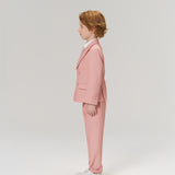 A young boy with light brown hair stands in profile, wearing the KIKI and JOJO Blush Reverie Suit Jacket over a white shirt with white sneakers, set against a plain light background.