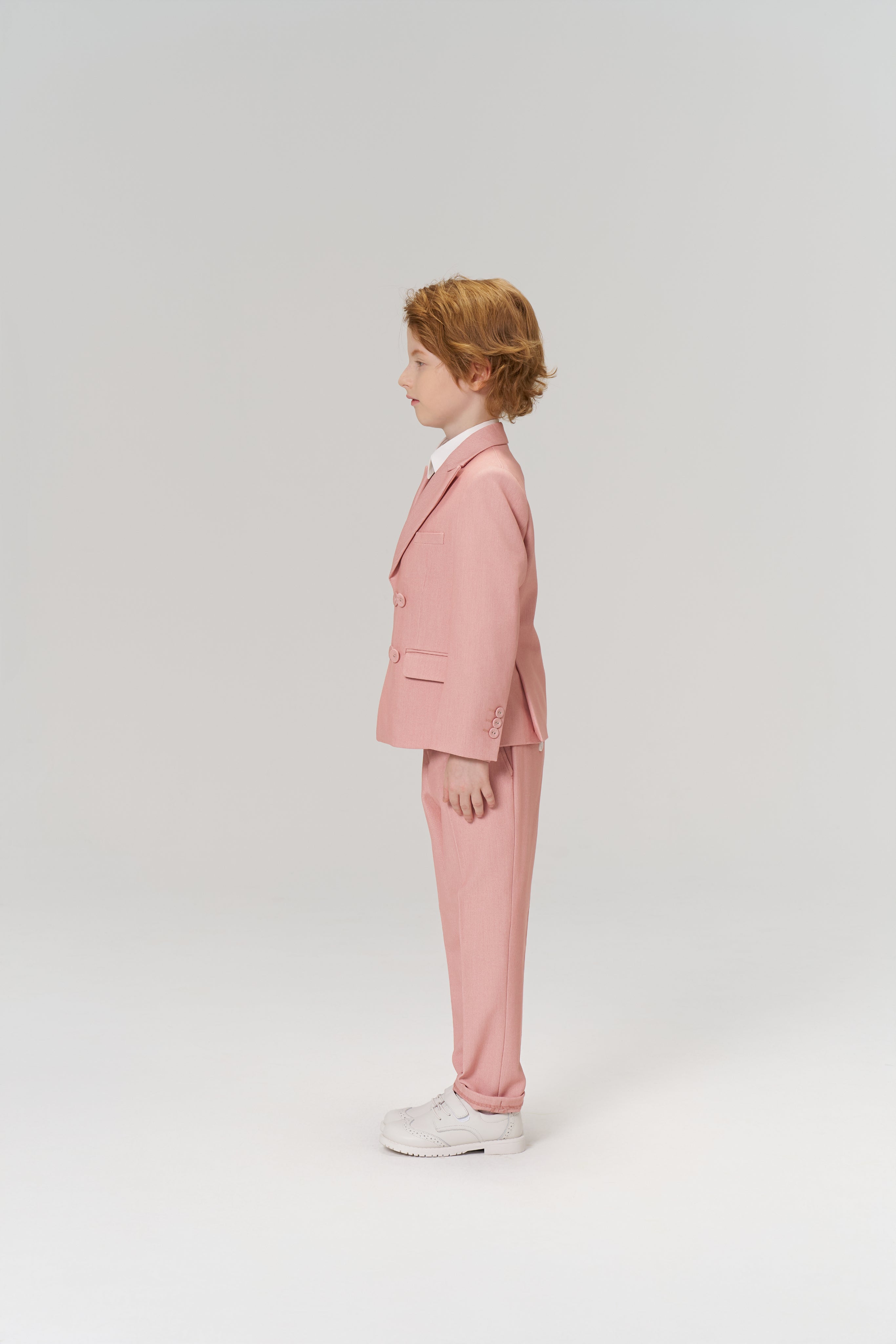 A young boy with light brown hair stands in profile, wearing the KIKI and JOJO Blush Reverie Suit Jacket over a white shirt with white sneakers, set against a plain light background.