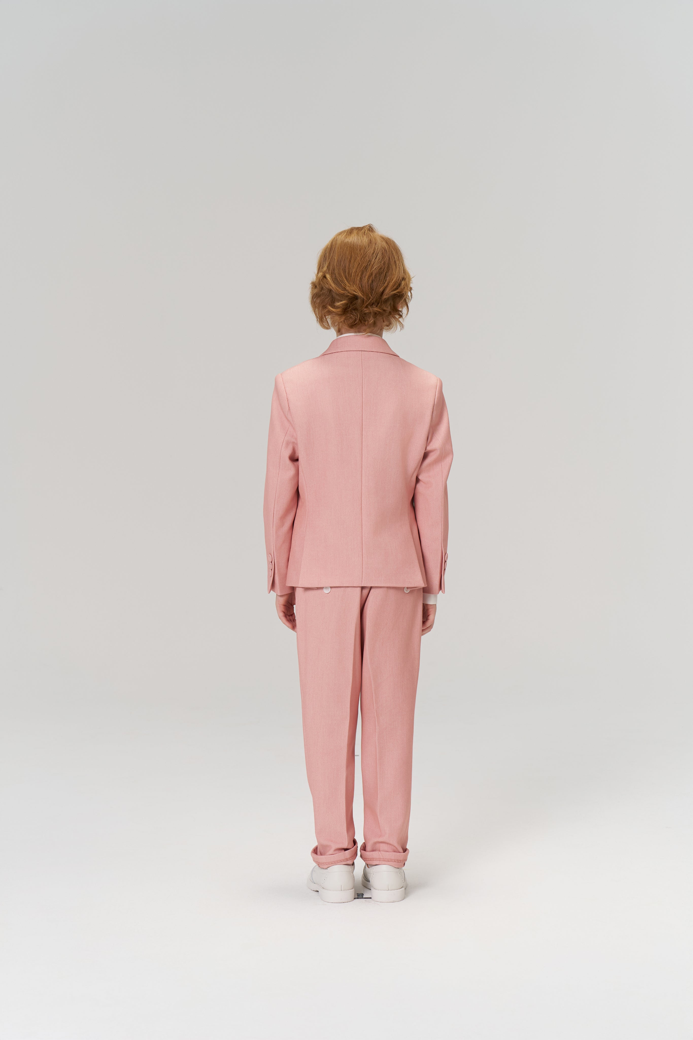 A child with short, light brown hair wears KIKI and JOJO Blush Reverie Suit Pants featuring an adjustable waistband, paired with white sneakers, standing facing away against a plain light background.