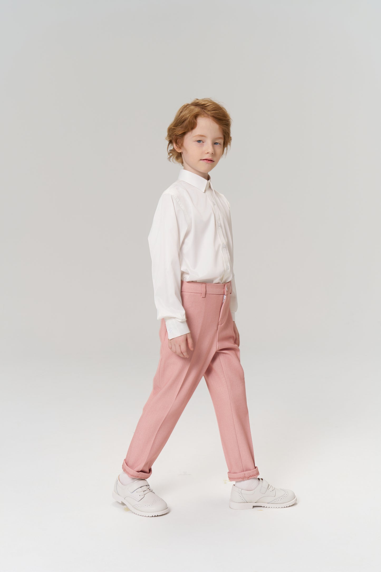 A young child with light brown hair stands against a plain background, wearing a white shirt, KIKI and JOJO Blush Reverie Suit Pants with an adjustable waistband, and white sneakers.