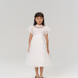 A young girl with long dark hair models the Whisper Bloom Dress by KIKI and JOJO, featuring embroidered floral details and bead accents, paired with silver shoes, as she stands facing forward against a plain white background.