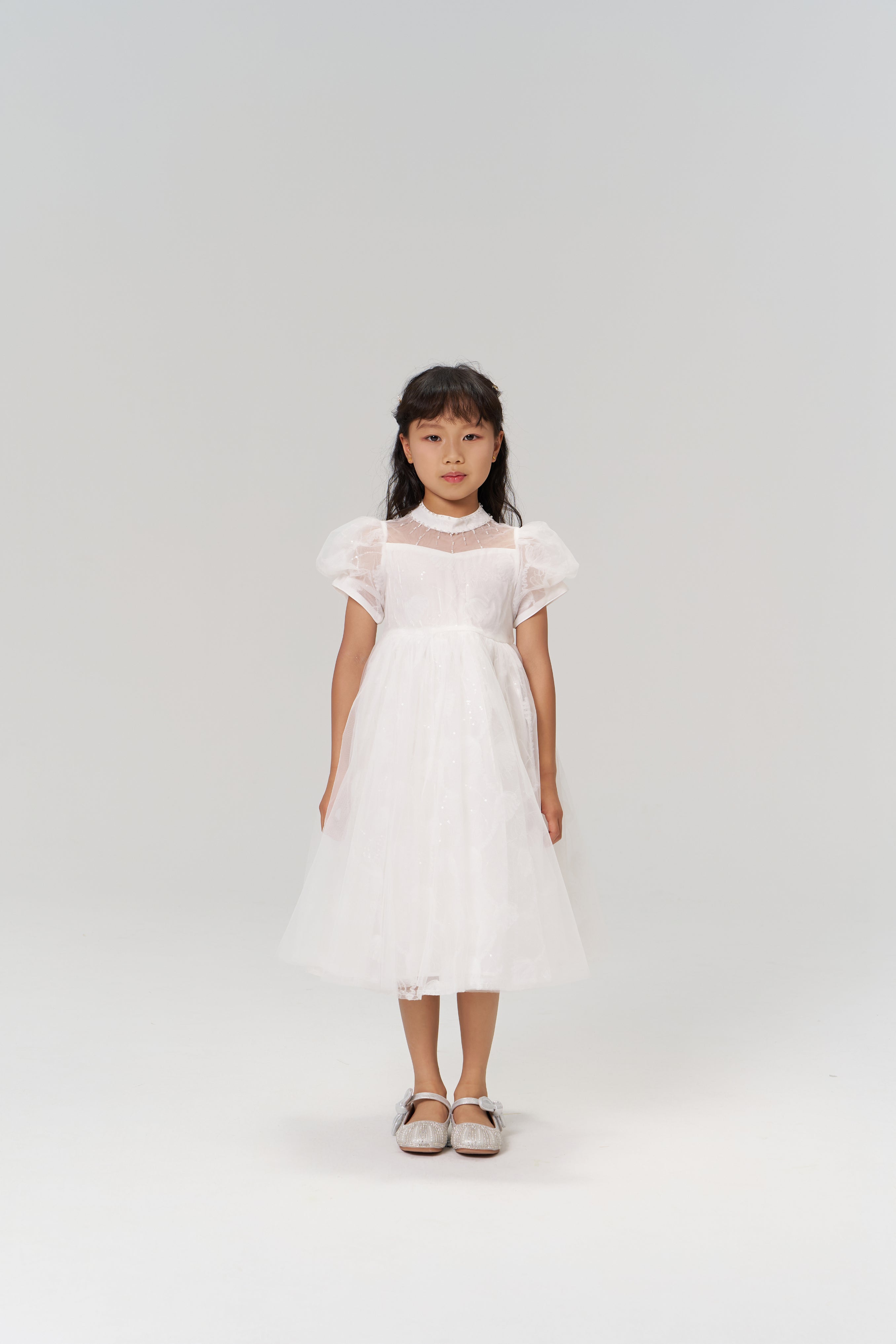 A young girl with long dark hair models the Whisper Bloom Dress by KIKI and JOJO, featuring embroidered floral details and bead accents, paired with silver shoes, as she stands facing forward against a plain white background.