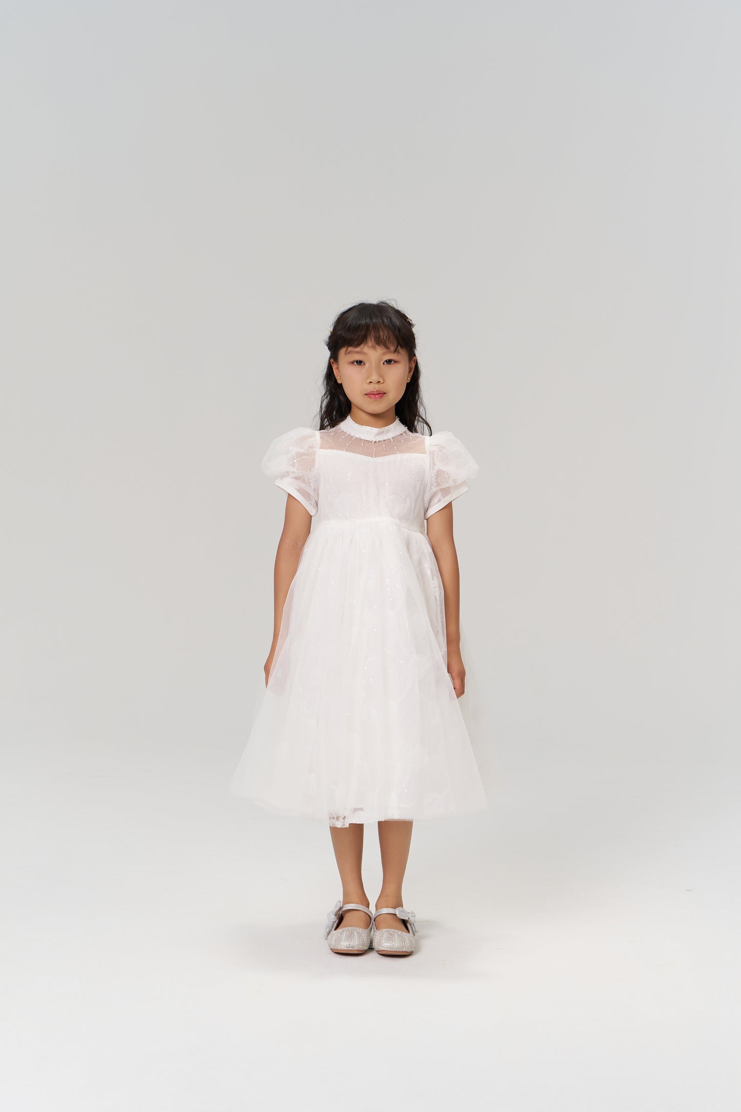 A young girl with long dark hair models the Whisper Bloom Dress by KIKI and JOJO, featuring embroidered floral details and bead accents, paired with silver shoes, as she stands facing forward against a plain white background.