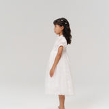 A young girl in the Whisper Bloom Dress by KIKI and JOJO, featuring embroidered florals and puff sleeves, stands in profile against a plain light background, paired with silver shoes.