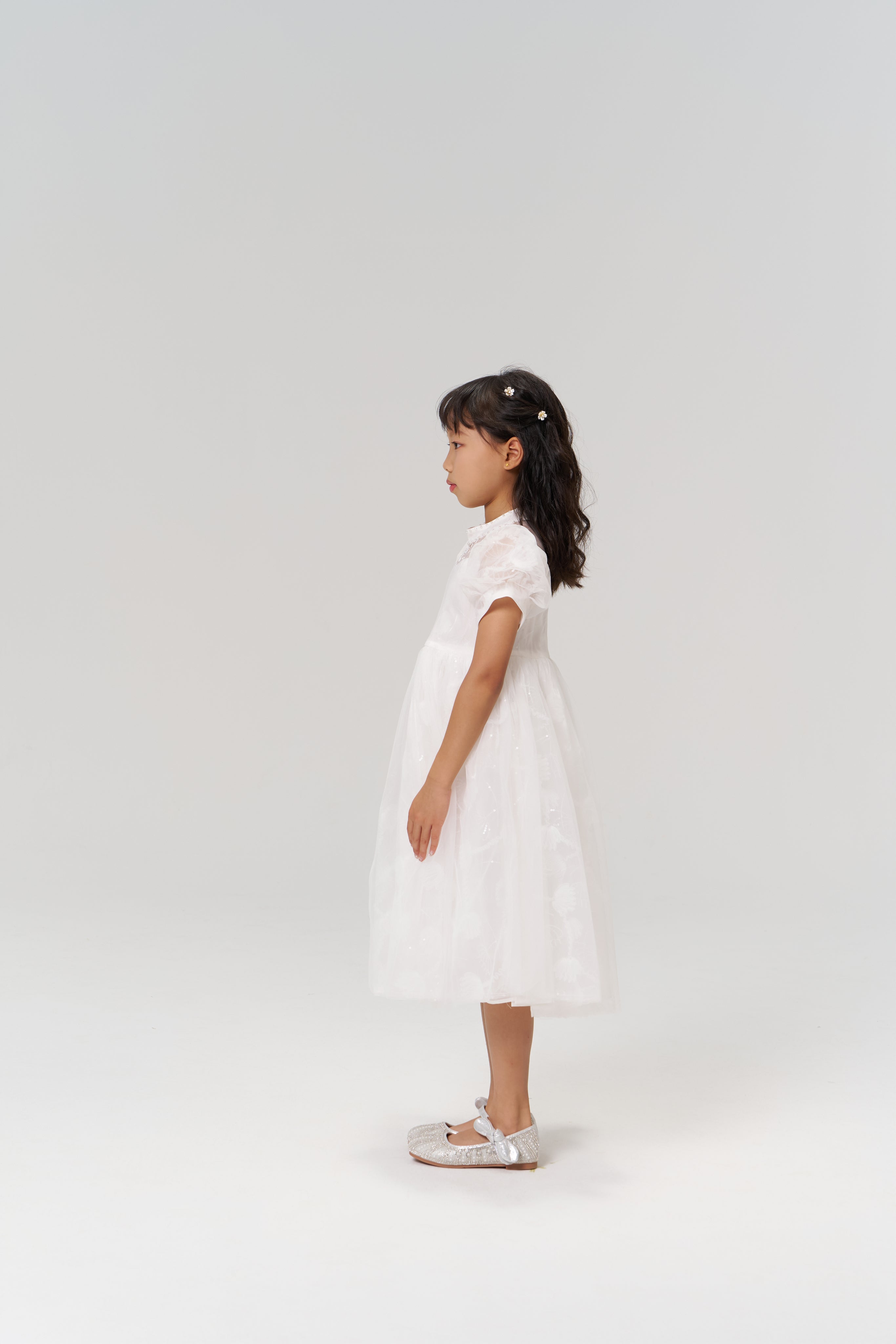 A young girl in the Whisper Bloom Dress by KIKI and JOJO, featuring embroidered florals and puff sleeves, stands in profile against a plain light background, paired with silver shoes.