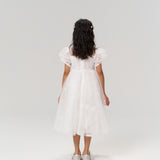A young girl stands facing away against a plain, light background, wearing the KIKI and JOJO Whisper Bloom Dress—a floral embroidered mini dress with puff sleeves—and silver shoes.