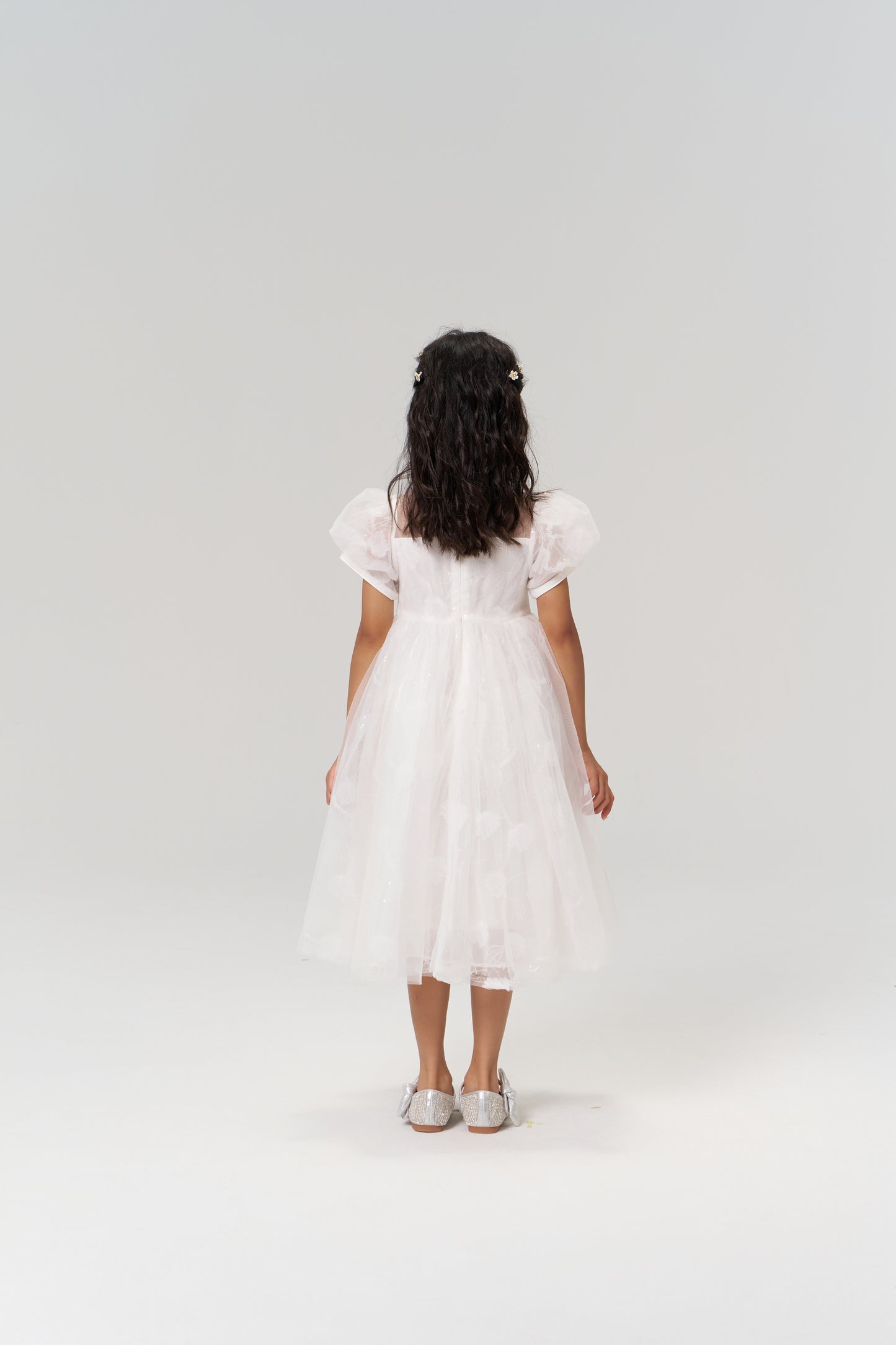 A young girl stands facing away against a plain, light background, wearing the KIKI and JOJO Whisper Bloom Dress—a floral embroidered mini dress with puff sleeves—and silver shoes.