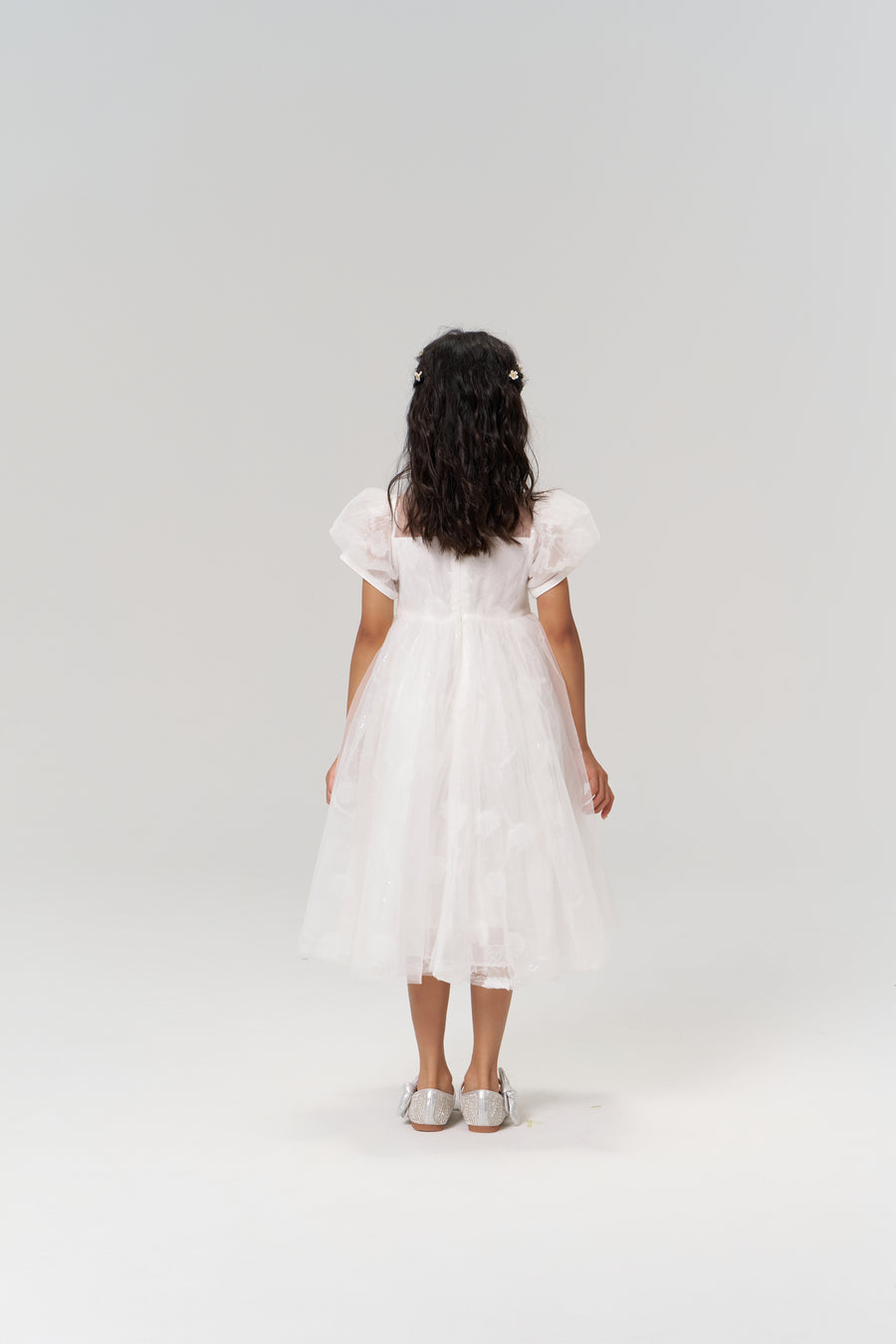 A young girl stands facing away against a plain, light background, wearing the KIKI and JOJO Whisper Bloom Dress—a floral embroidered mini dress with puff sleeves—and silver shoes.