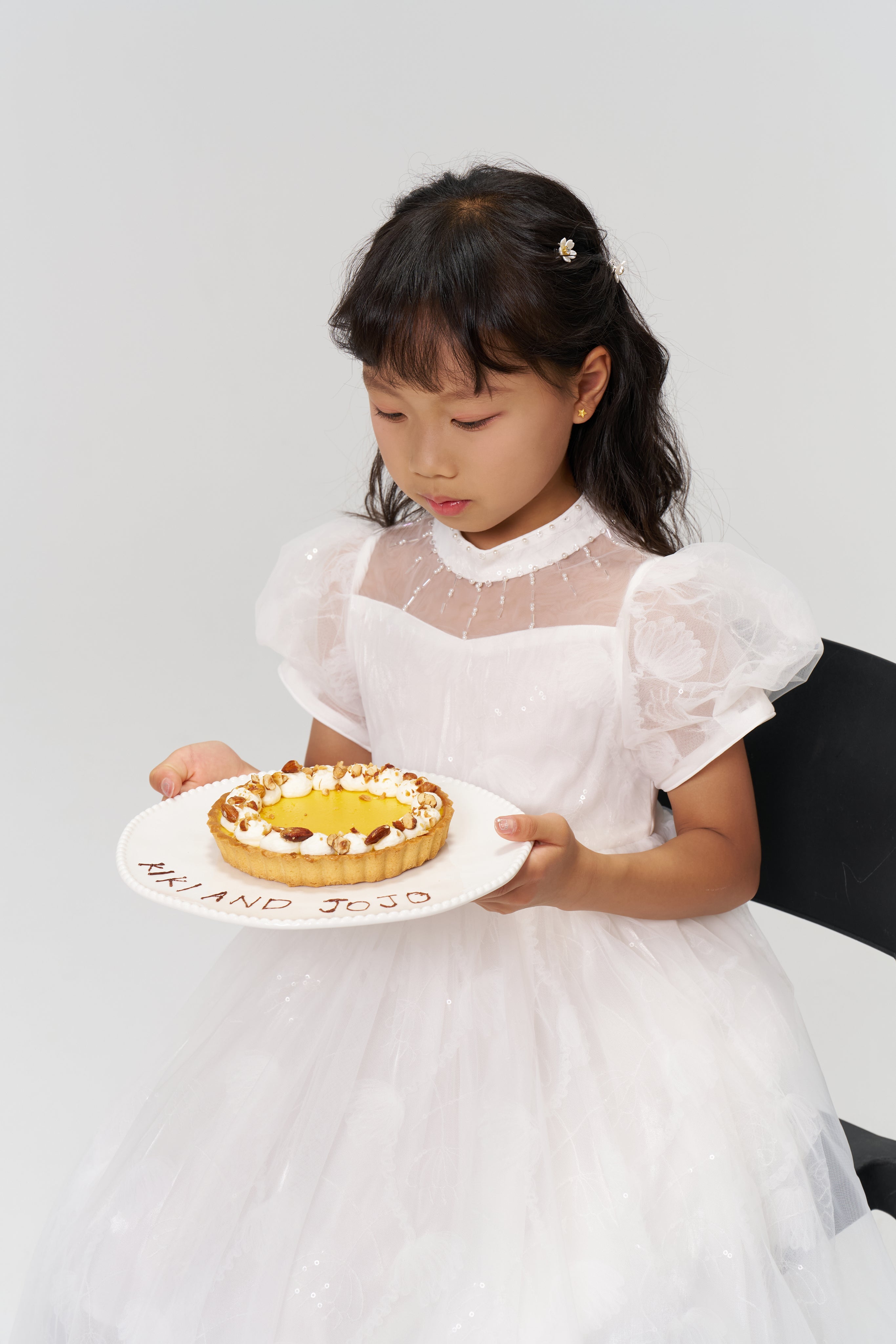 A young girl in a white dress sits on a black chair, holding a plate with a decorated cake. The plate has “HAPPY BDAY” written in icing.