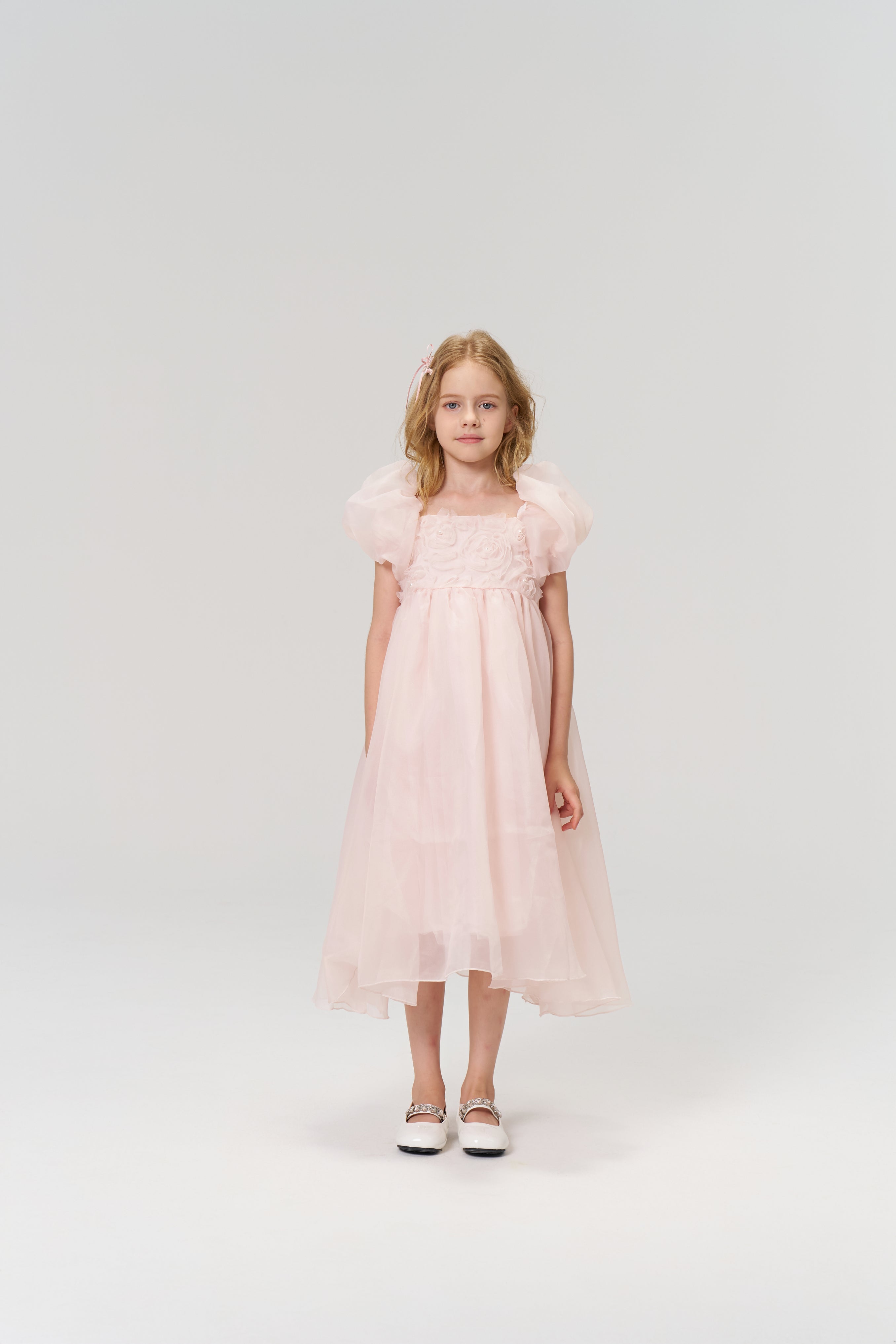 A young girl poses in a studio wearing the KIKI and JOJO Blush Whisper Dress, a princess-style blush pink outfit with puff sleeves, paired with white shoes.