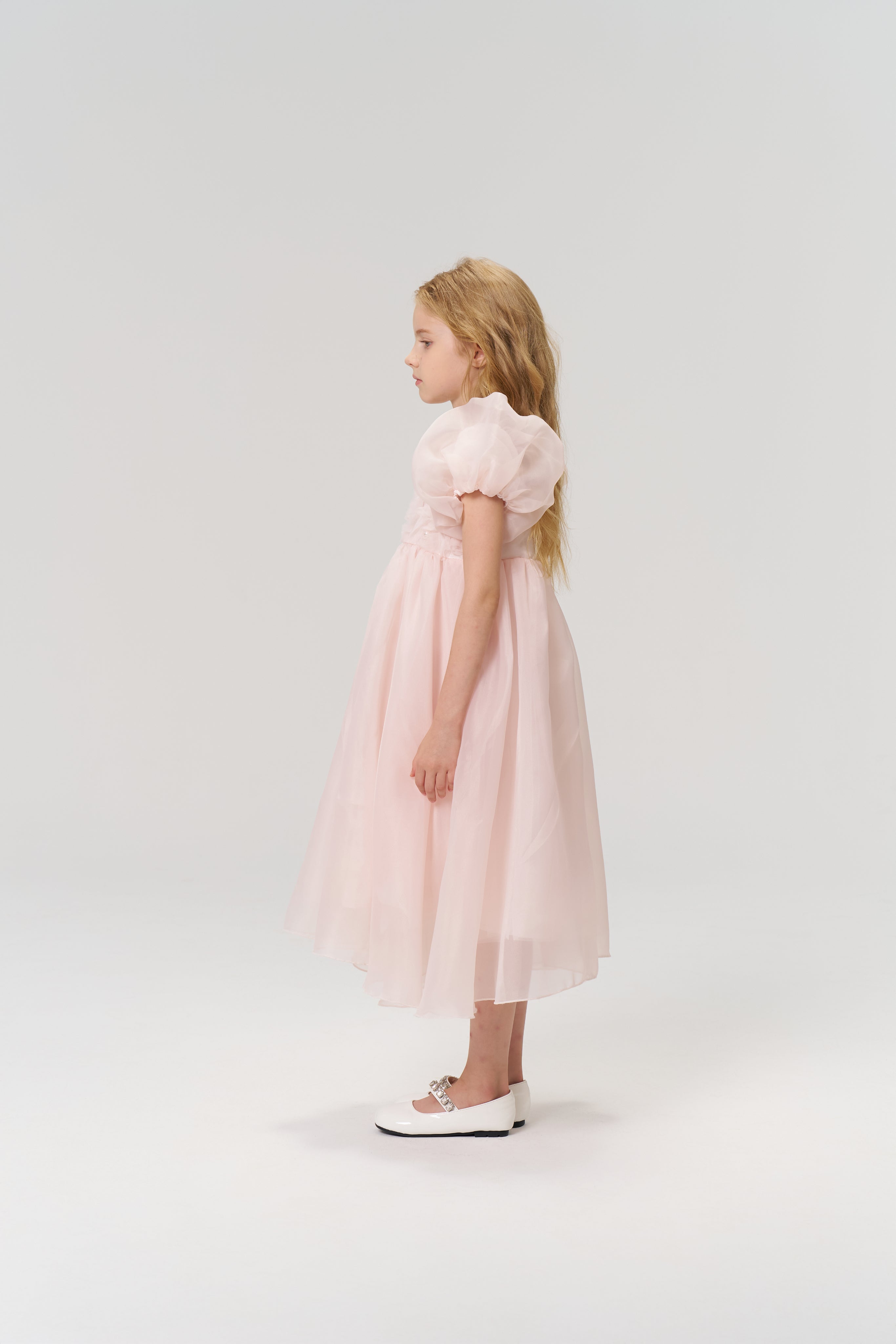 A young girl stands in profile against a plain light background, wearing the KIKI and JOJO Blush Whisper Dress—a princess-style blush pink dress with puff sleeves—and white shoes.
