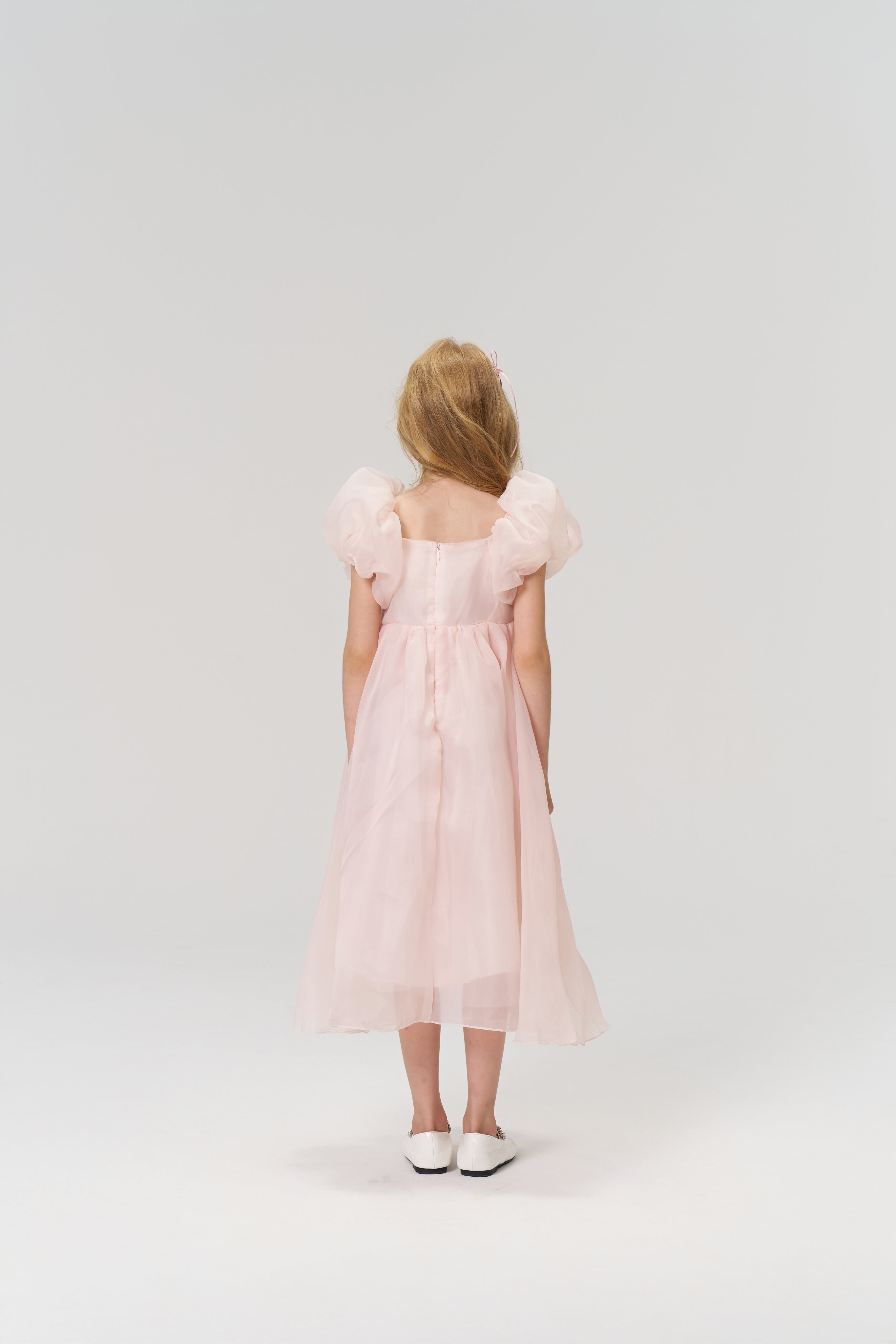 A young girl with light brown hair wears the KIKI and JOJO Blush Whisper Dress— a princess-style blush pink gown with puff sleeves and 3D floral appliqués—while standing against a plain, light background.