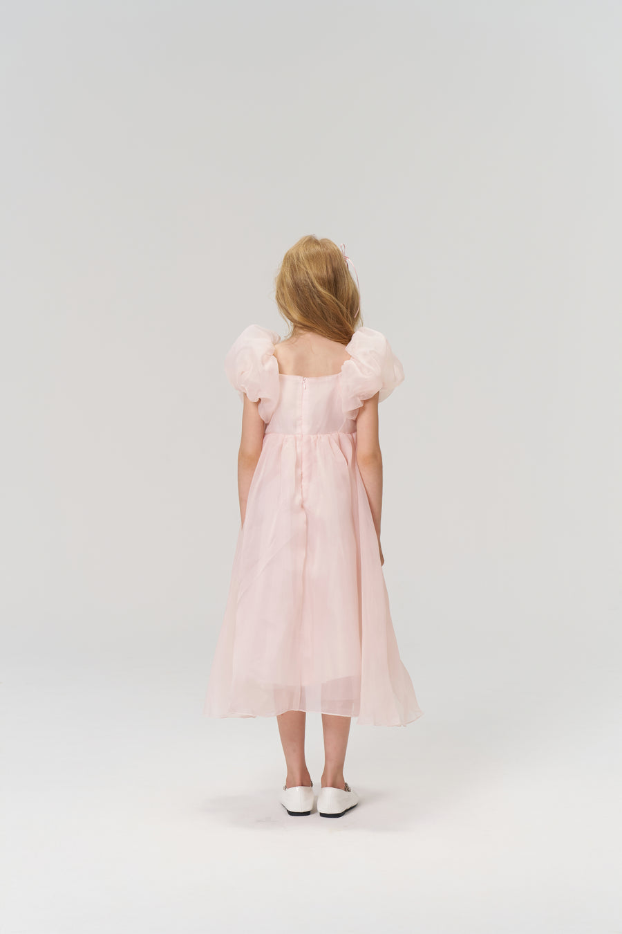 A young girl with light brown hair wears the KIKI and JOJO Blush Whisper Dress— a princess-style blush pink gown with puff sleeves and 3D floral appliqués—while standing against a plain, light background.
