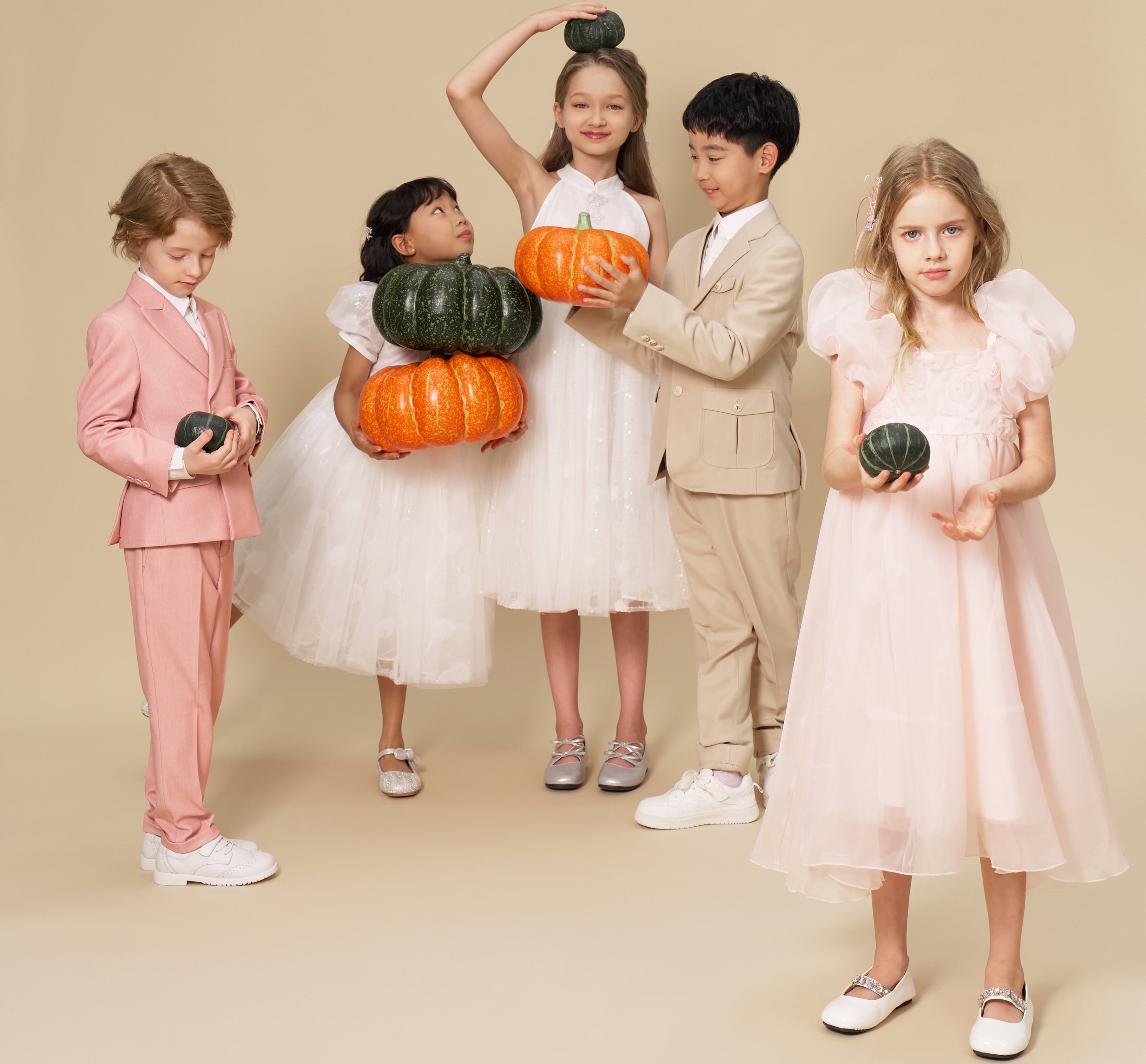 Five children in formal outfits stand together, some holding pumpkins of various sizes and colors, against a plain beige background.