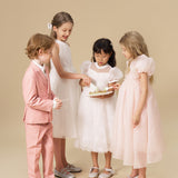Four children dressed in formal attire stand together; one girl holds a small cake while the others look at it. The background is plain beige.