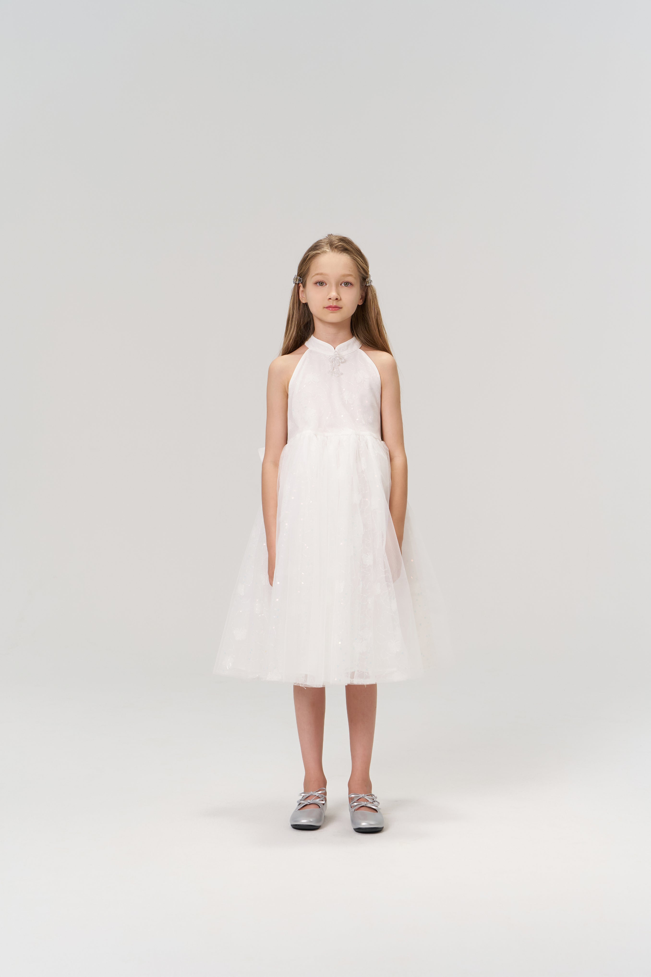 A young girl faces forward in the KIKI and JOJO Butterfly Whispers Dress—a sleeveless white halter dress with a layered tulle skirt—paired with silver shoes, set against a plain light background.