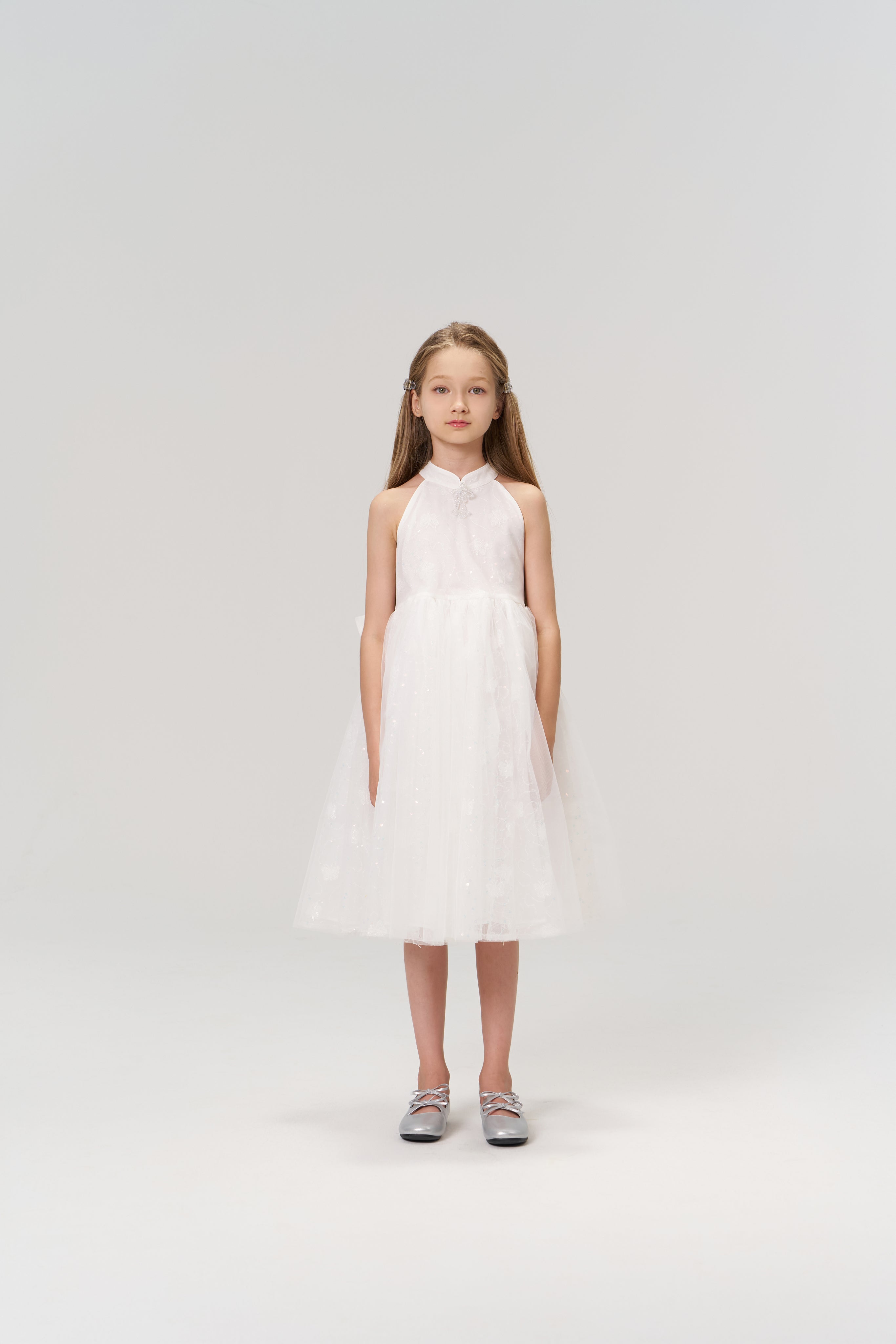 A young girl faces forward in the KIKI and JOJO Butterfly Whispers Dress—a sleeveless white halter dress with a layered tulle skirt—paired with silver shoes, set against a plain light background.