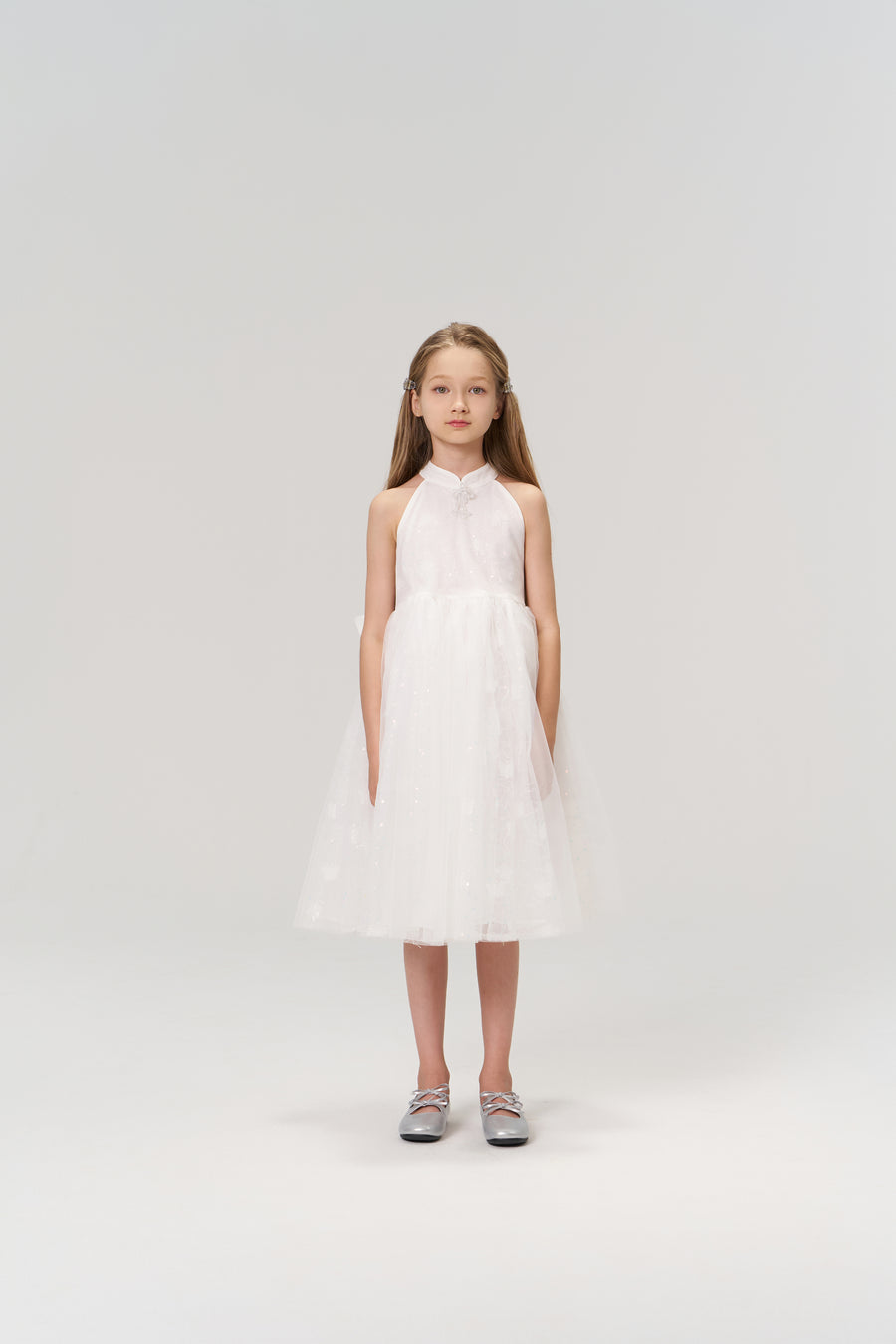 A young girl faces forward in the KIKI and JOJO Butterfly Whispers Dress—a sleeveless white halter dress with a layered tulle skirt—paired with silver shoes, set against a plain light background.