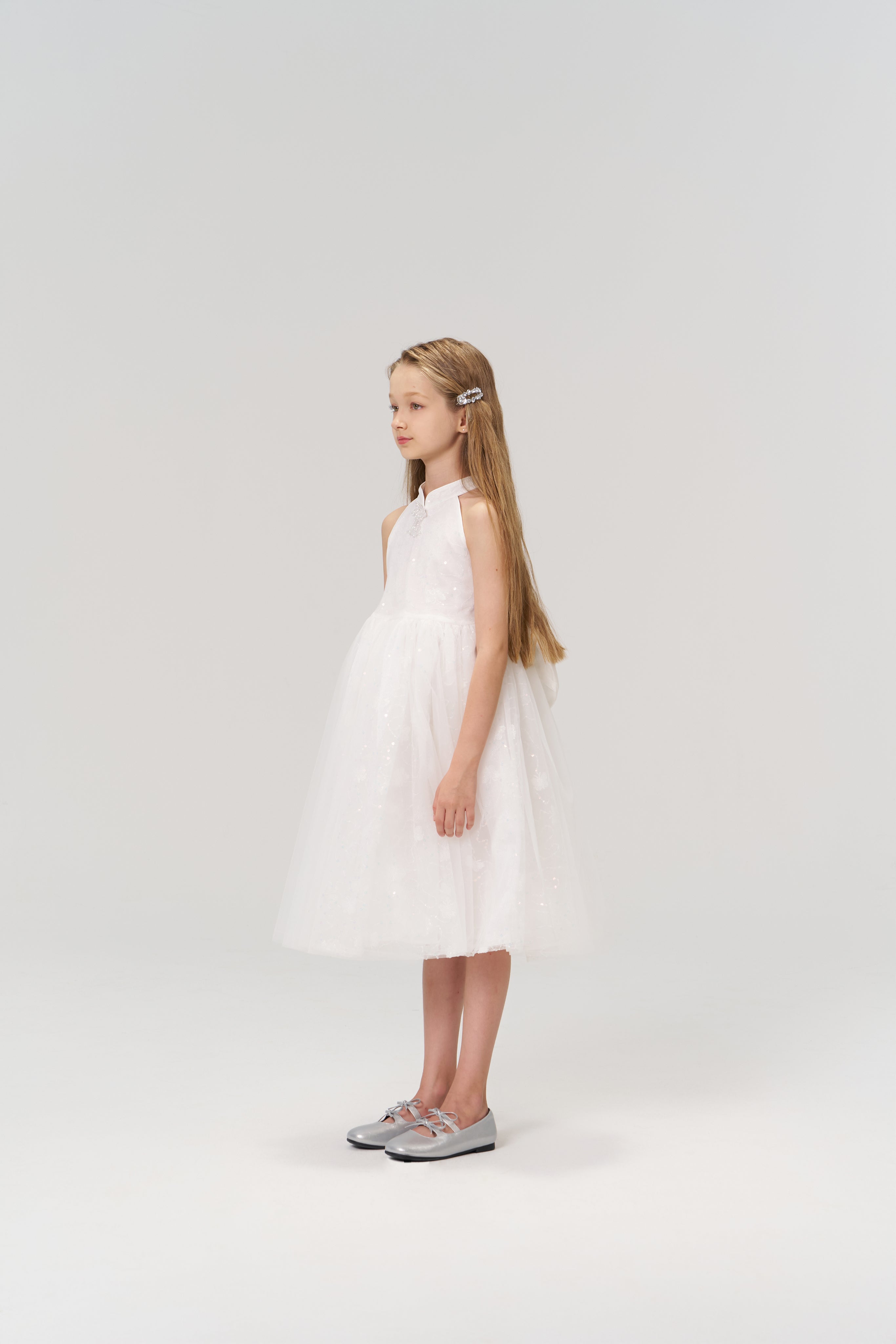 A young girl with long hair stands sideways in the KIKI and JOJO Butterfly Whispers Dress, paired with silver shoes, against a plain light background.